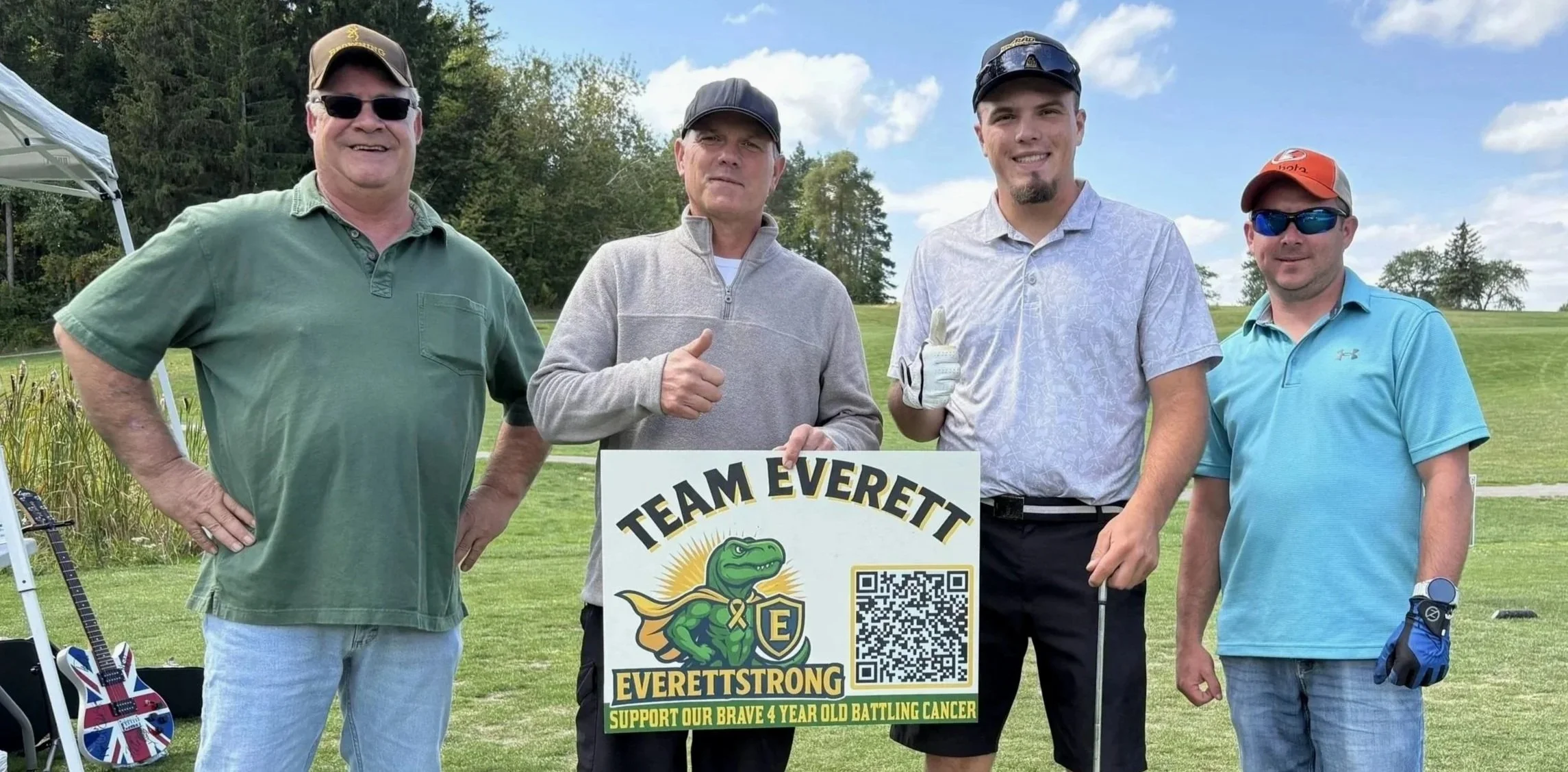 Four men standing outdoors on a golf course, holding a sign that reads 'TEAM EVERETT EVERETTSTRONG SUPPORT OUR BRAVE 4 YEAR OLD BATTLING CANCER.' The second man from the left is giving a thumbs-up. All men are dressed in casual golf attire and sport sunglasses or caps.
