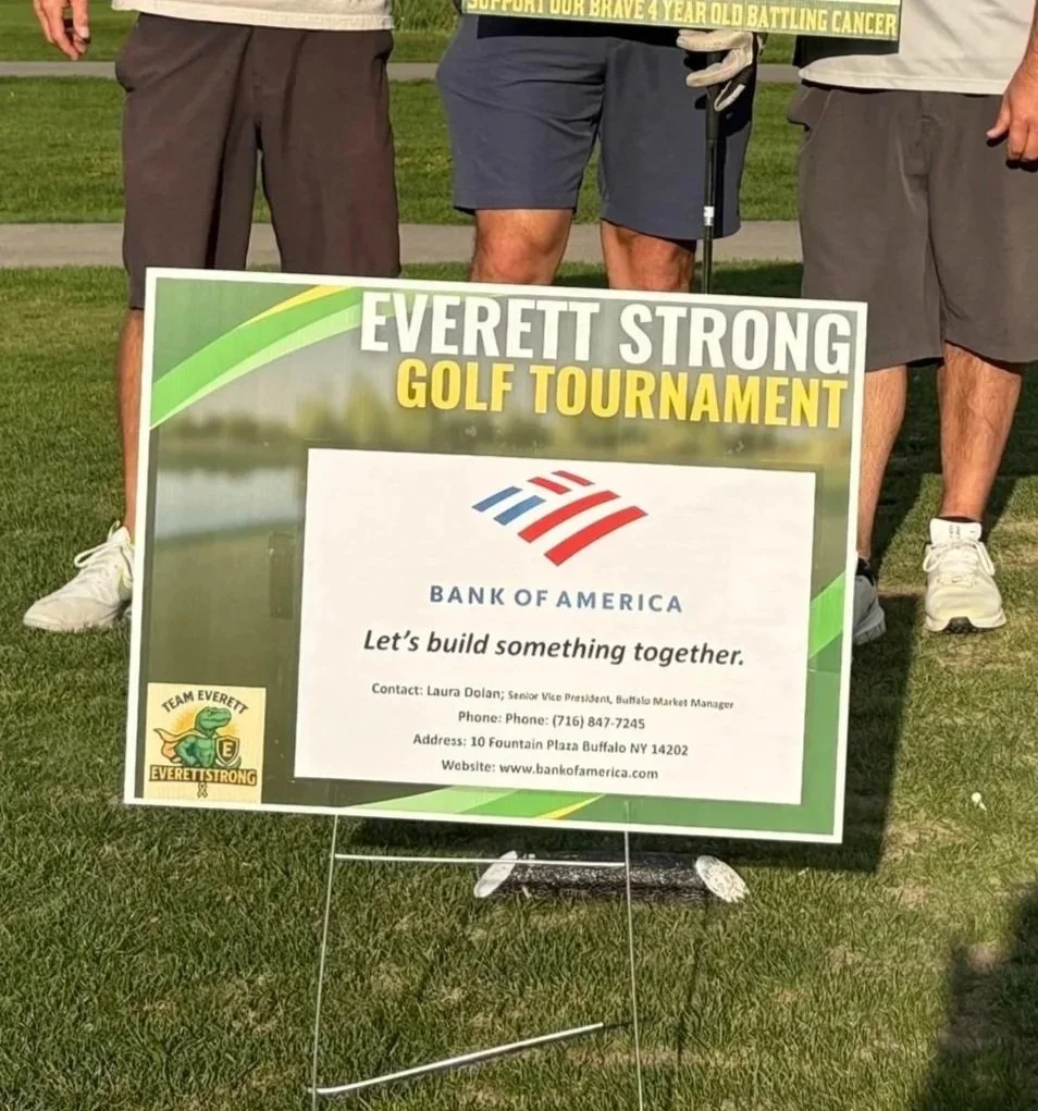 Sign at a golf tournament supporting Everett Strong and Bank of America, with three men partially visible standing behind it on a grassy field.