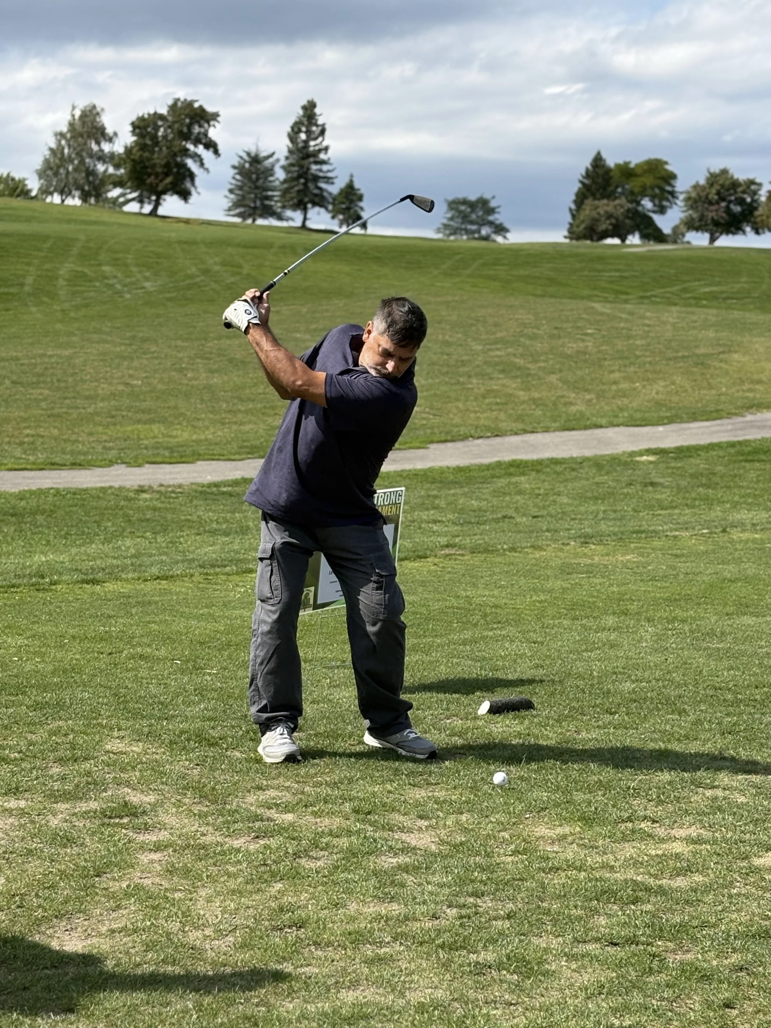 A man in a dark shirt and gray shorts practicing golf on a lush, green golf course, swinging a club near a golf ball with a scenic background of trees and cloudy sky.