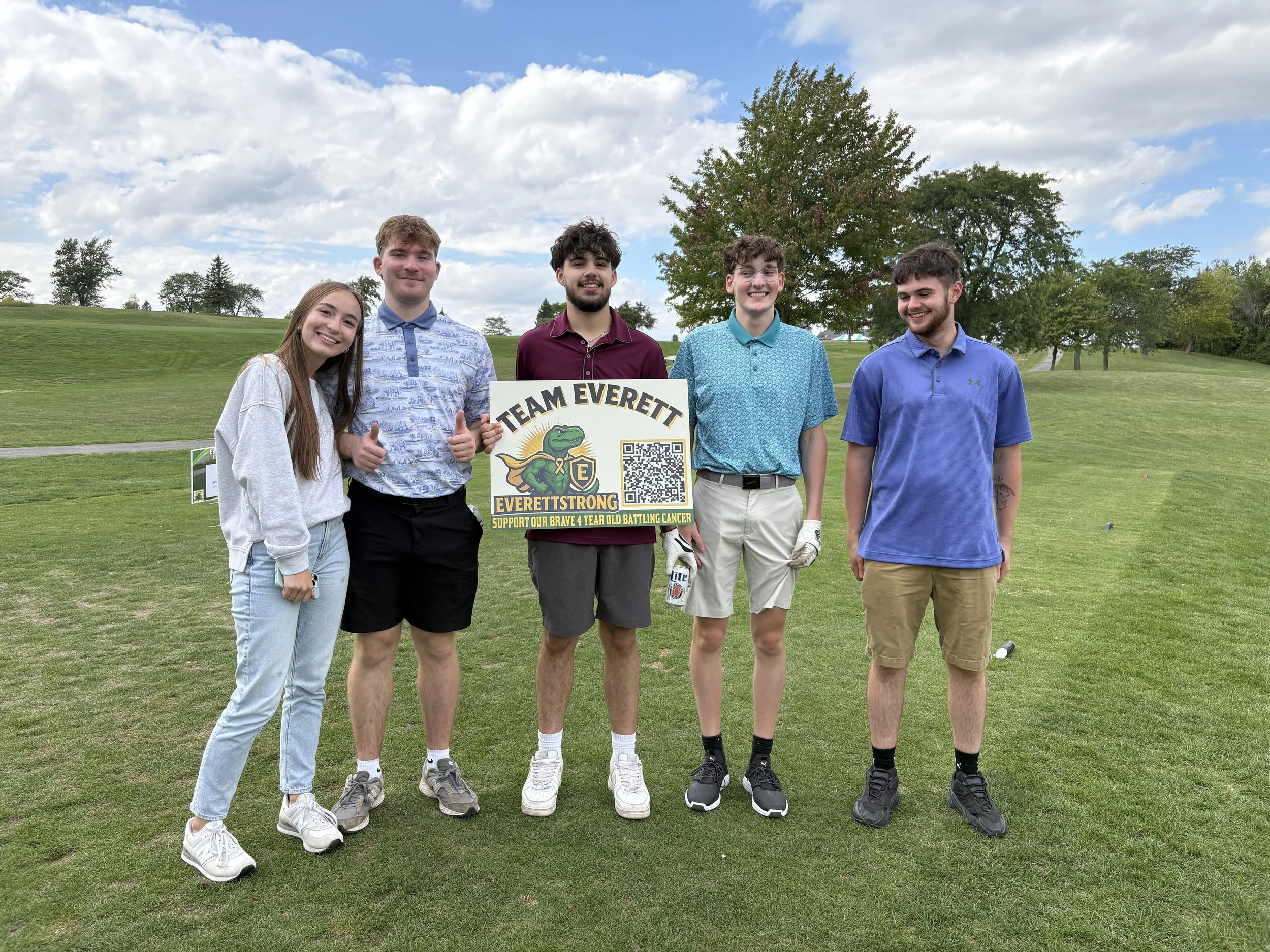 Group of five young people standing on a golf course, holding a sign that says 'Team Everett' and support for a four-year-old battling cancer, smiling and dressed casually.
