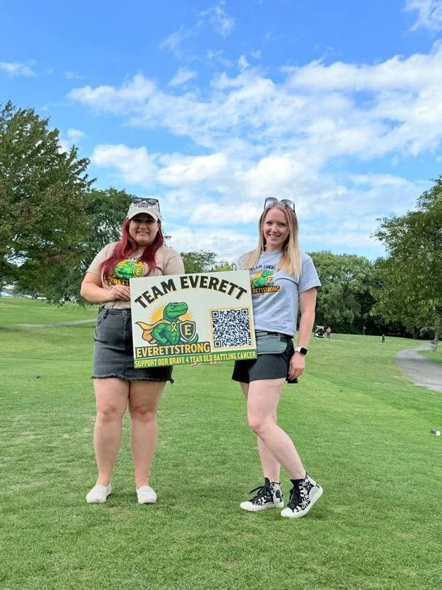 Two women standing on a golf course holding a yellow sign that reads 'Team Everett' and 'EverettStrong' with a QR code and a dinosaur logo, participating in a cancer support event.
