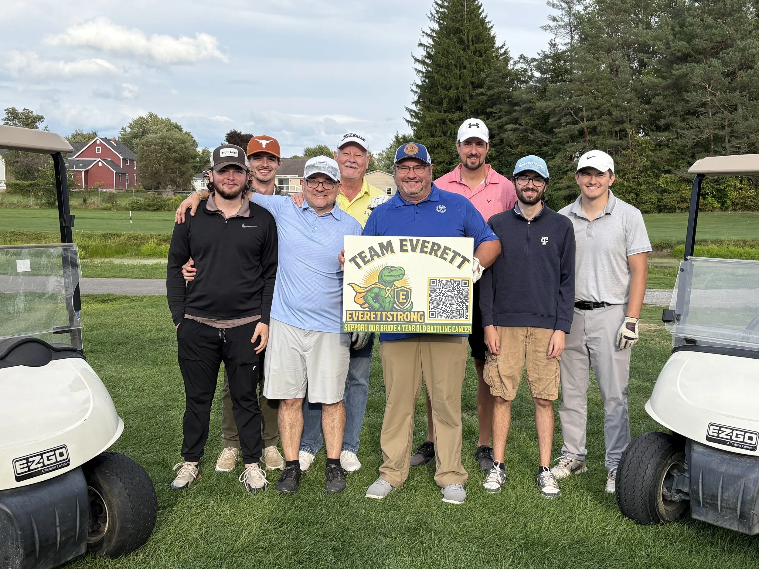 Group of eight men standing on a golf course holding a sign that reads 'TEAM EVEREST' with a cartoon mountain climber logo and QR code, surrounded by golf carts and trees.