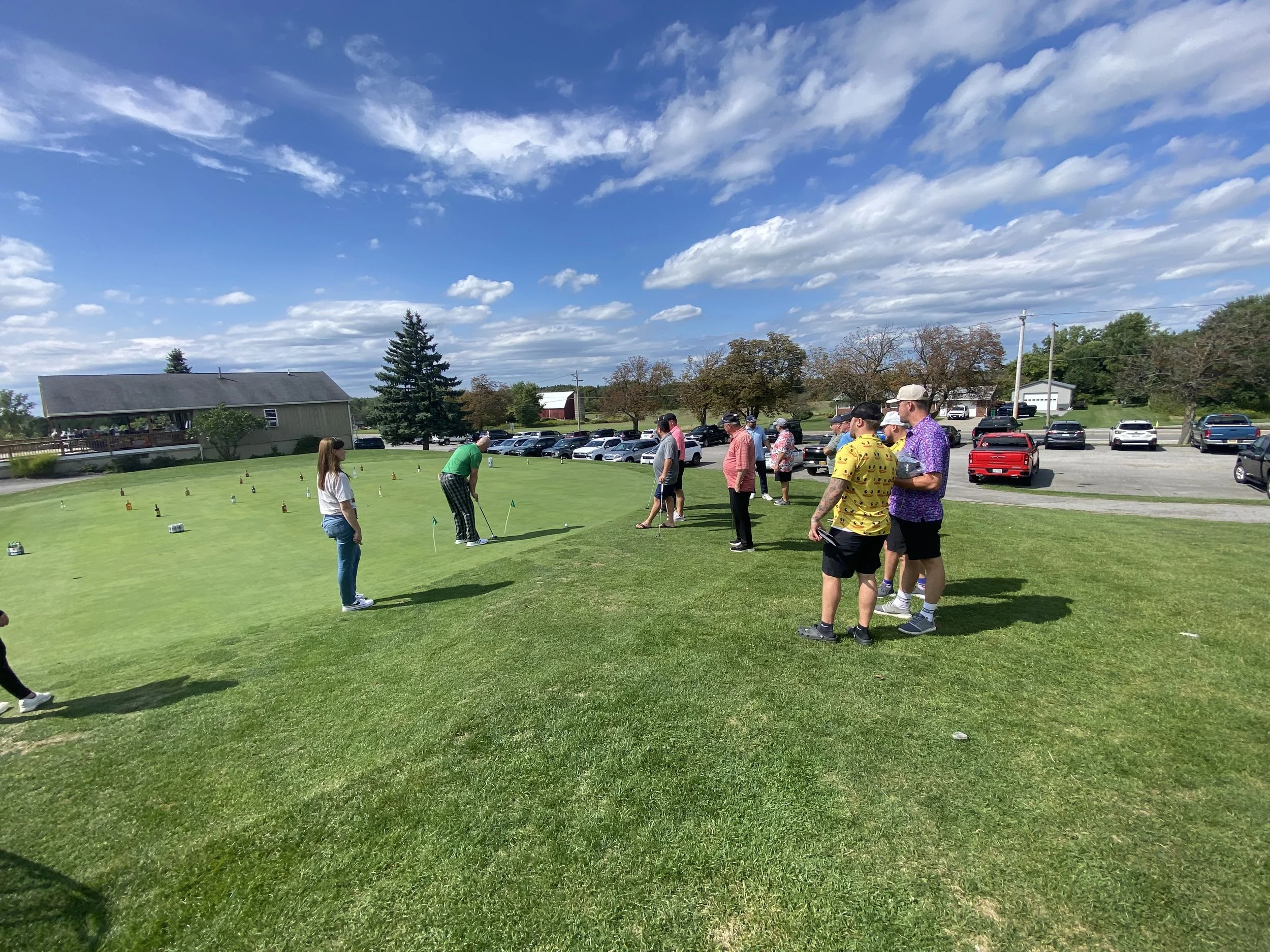 A group of people playing or practicing mini golf outdoors on a grass green with golf clubs in hand. The sky is partly cloudy with a clear blue sky. In the background, there is a parking lot filled with cars, a barn, some trees, and a building with a
