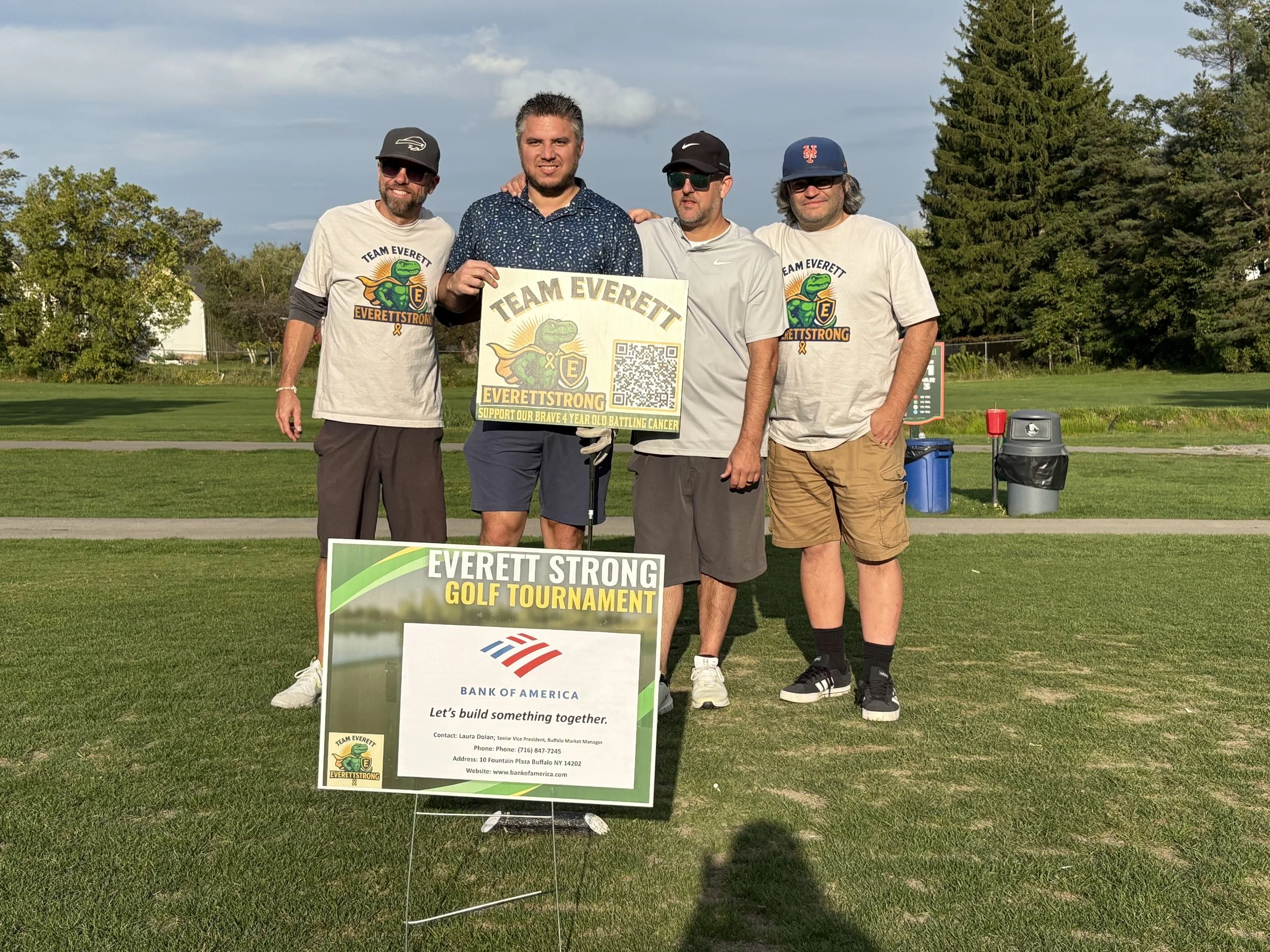 Four men standing on a golf course holding a sign that reads "Team Everett Everrett Strong Golf Tournament". They are wearing casual clothing and sunglasses. Behind them, there are trees, a blue sky with clouds, and a few golf-related items like a tr