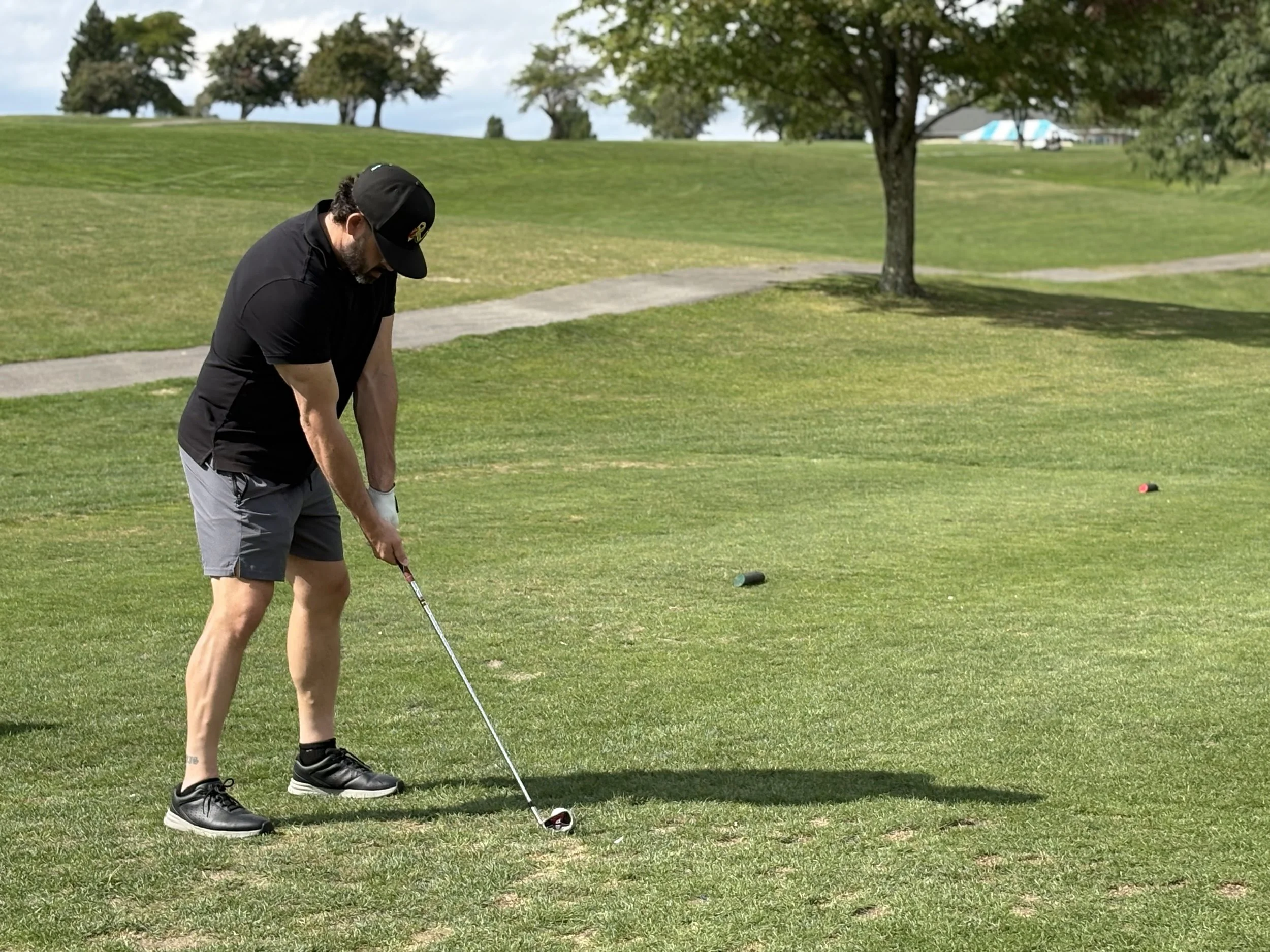 A man wearing a black cap, black shirt, gray shorts, and black shoes is playing golf on a green golf course. He is preparing to hit the golf ball with a club, with a golf tee visible on the ground. There are trees and a small path in the background u