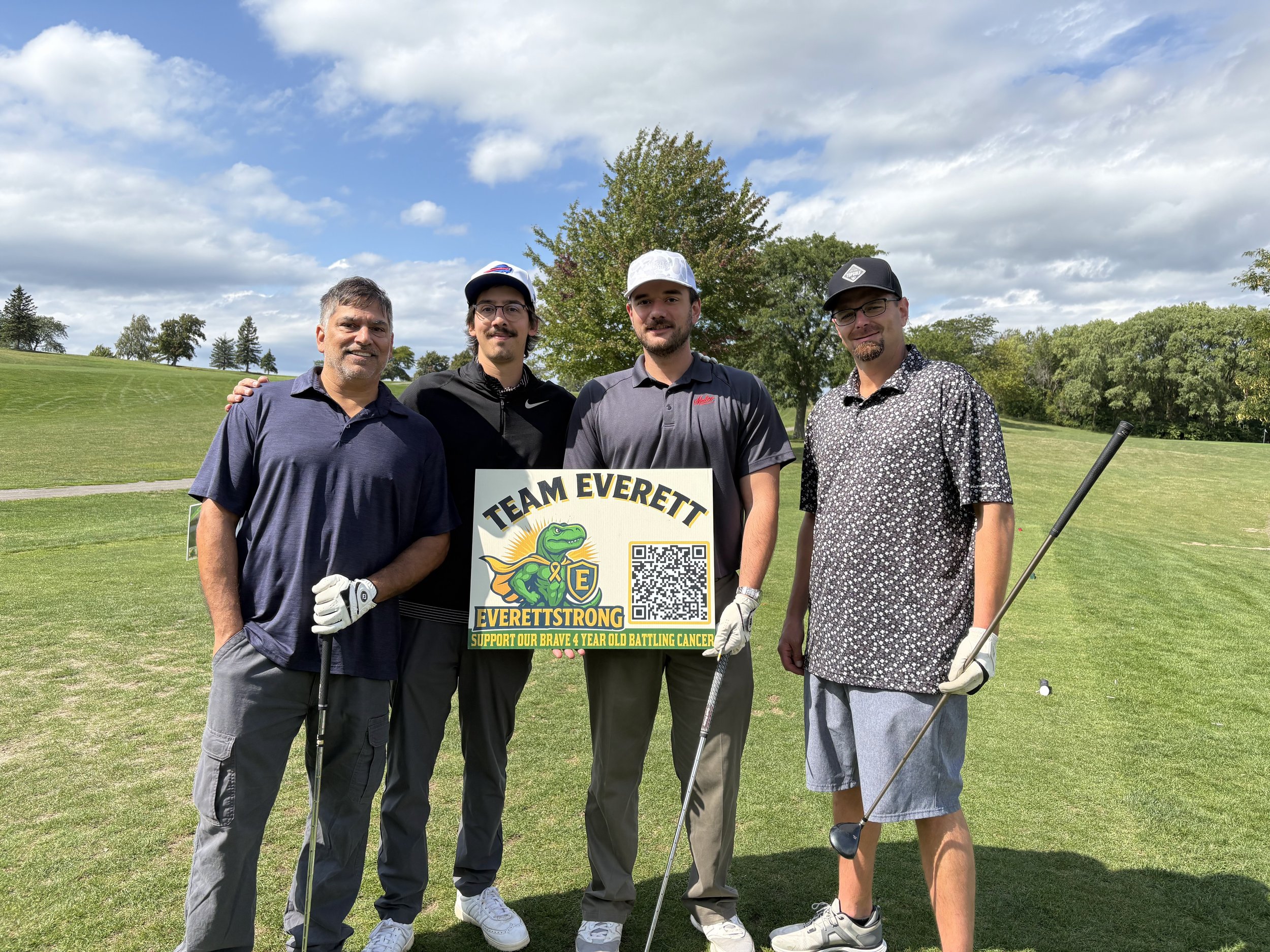 Four men standing on a golf course posing for a photo. They are holding a sign supporting a young boy battling cancer. The background shows a green landscape with trees and a partly cloudy sky.