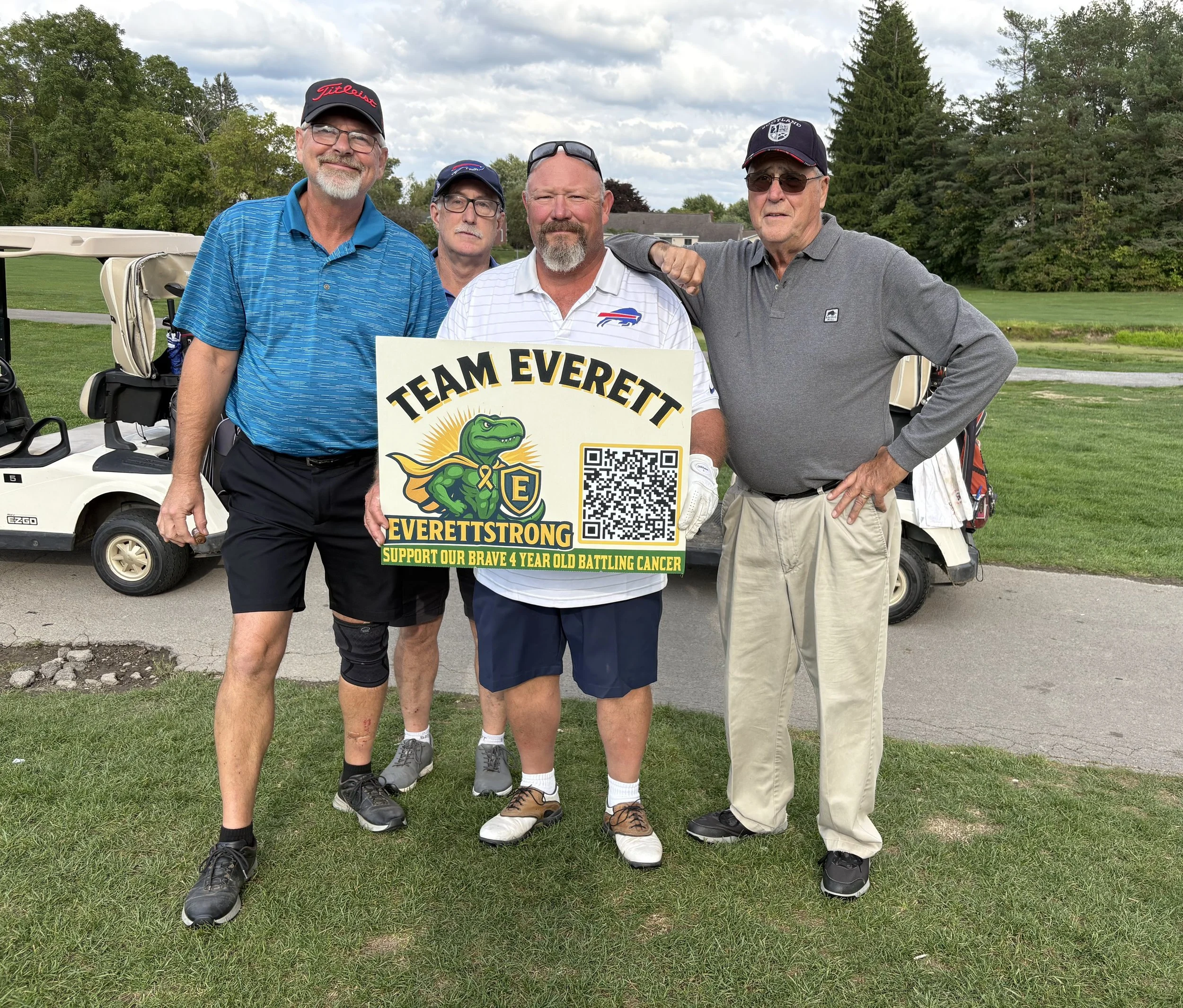 Four men standing on a golf course holding a sign supporting children with cancer. They are dressed in golf attire and are standing in front of a golf cart with trees and a house in the background.