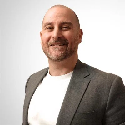 Portrait of a smiling man wearing a gray blazer and white t-shirt against a plain white background.