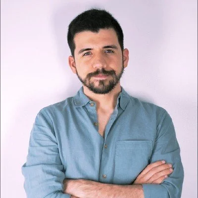 Carlos Melo, standing with his arms crossed in-front of a white background with a blue shirt.