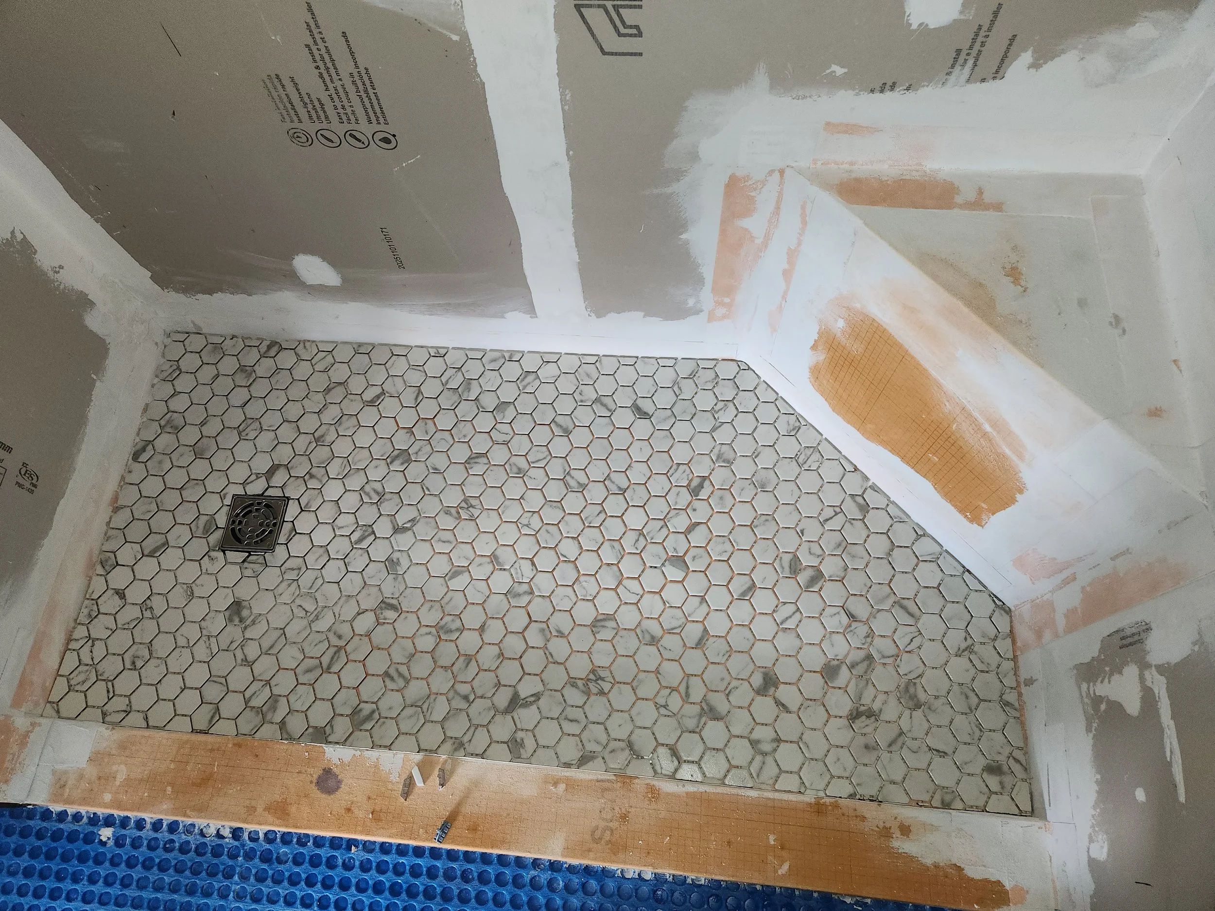 A bathroom under renovation with hexagonal tile flooring, partially installed wall drywall, and a drain on the floor.