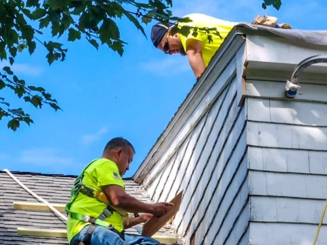 Two workers in yellow shirts working on the roof of a house, one above and one below, during daylight.
