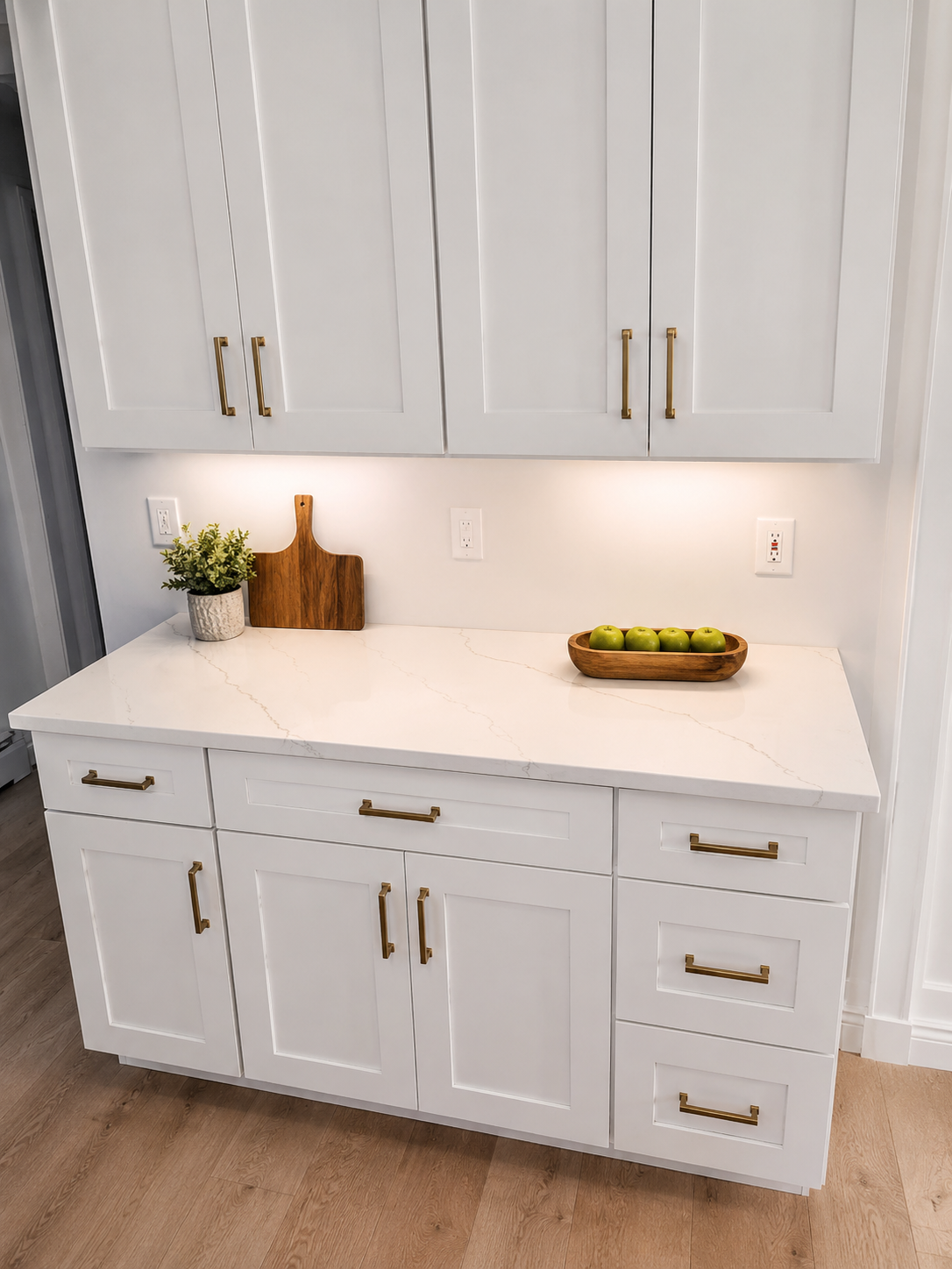 White kitchen countertop with gold drawer handles, a potted plant, a wooden cutting board, a wooden bowl with green apples, and white cabinets with gold handles.