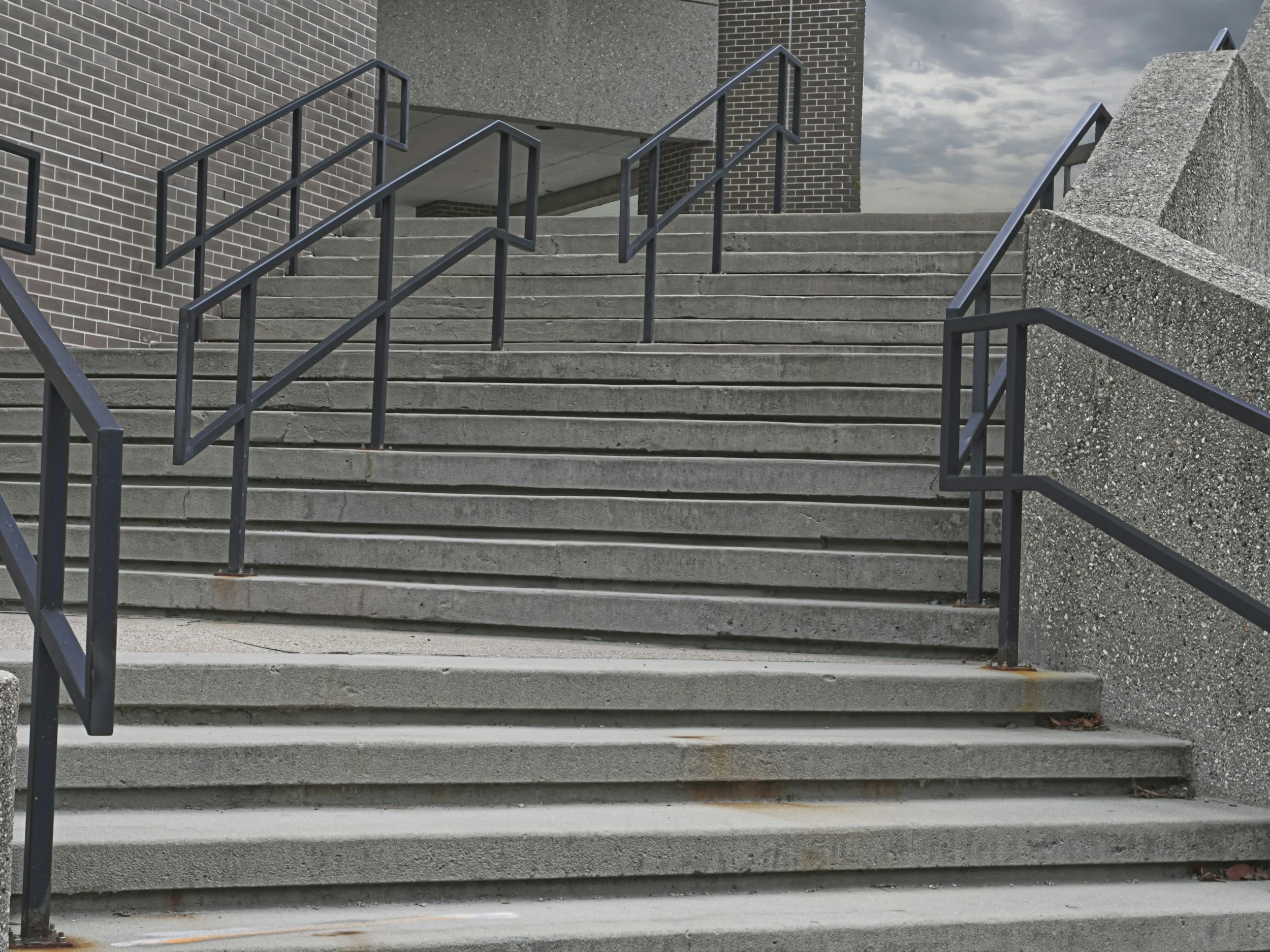 Concrete outdoor stairs with black metal handrails leading up to a building with brick and concrete walls, under a cloudy sky.