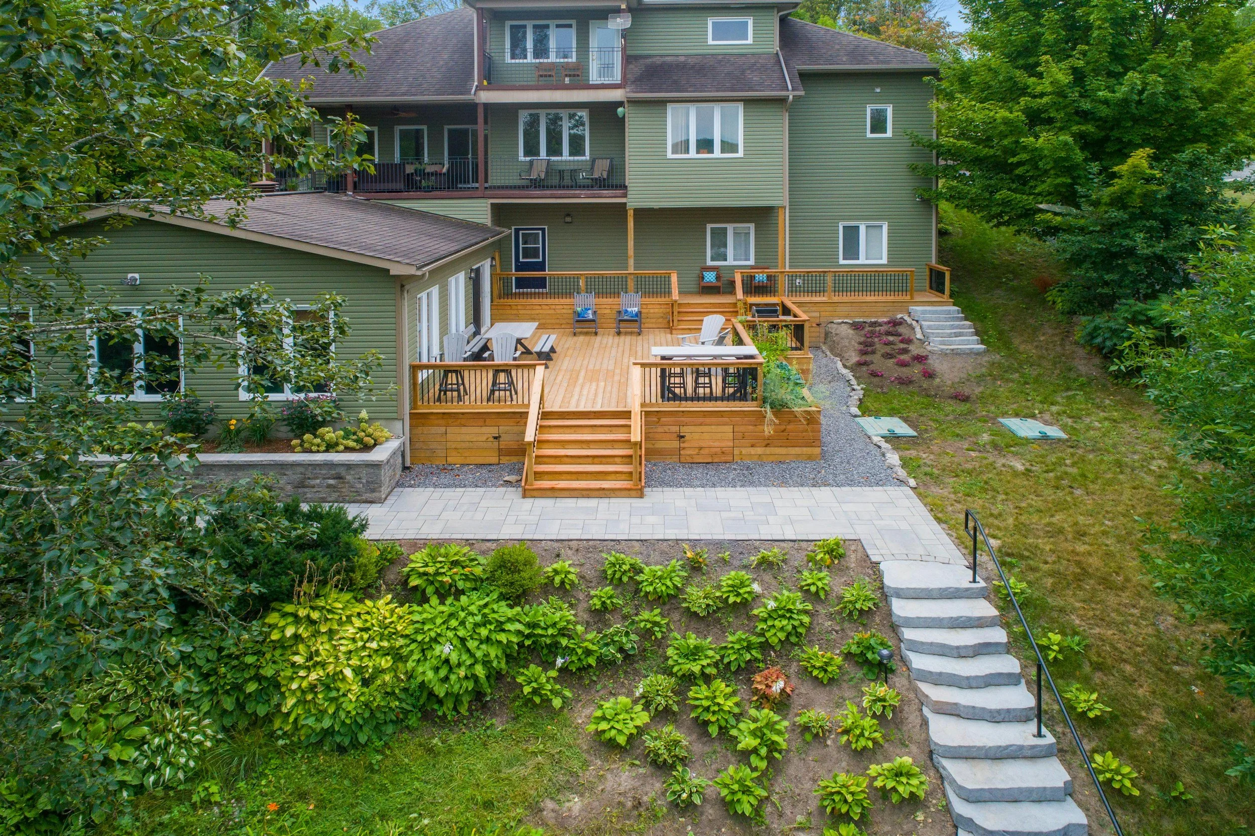 A green multi-story house with a large wooden deck and an outdoor dining area, surrounded by lush greenery and landscaped garden.