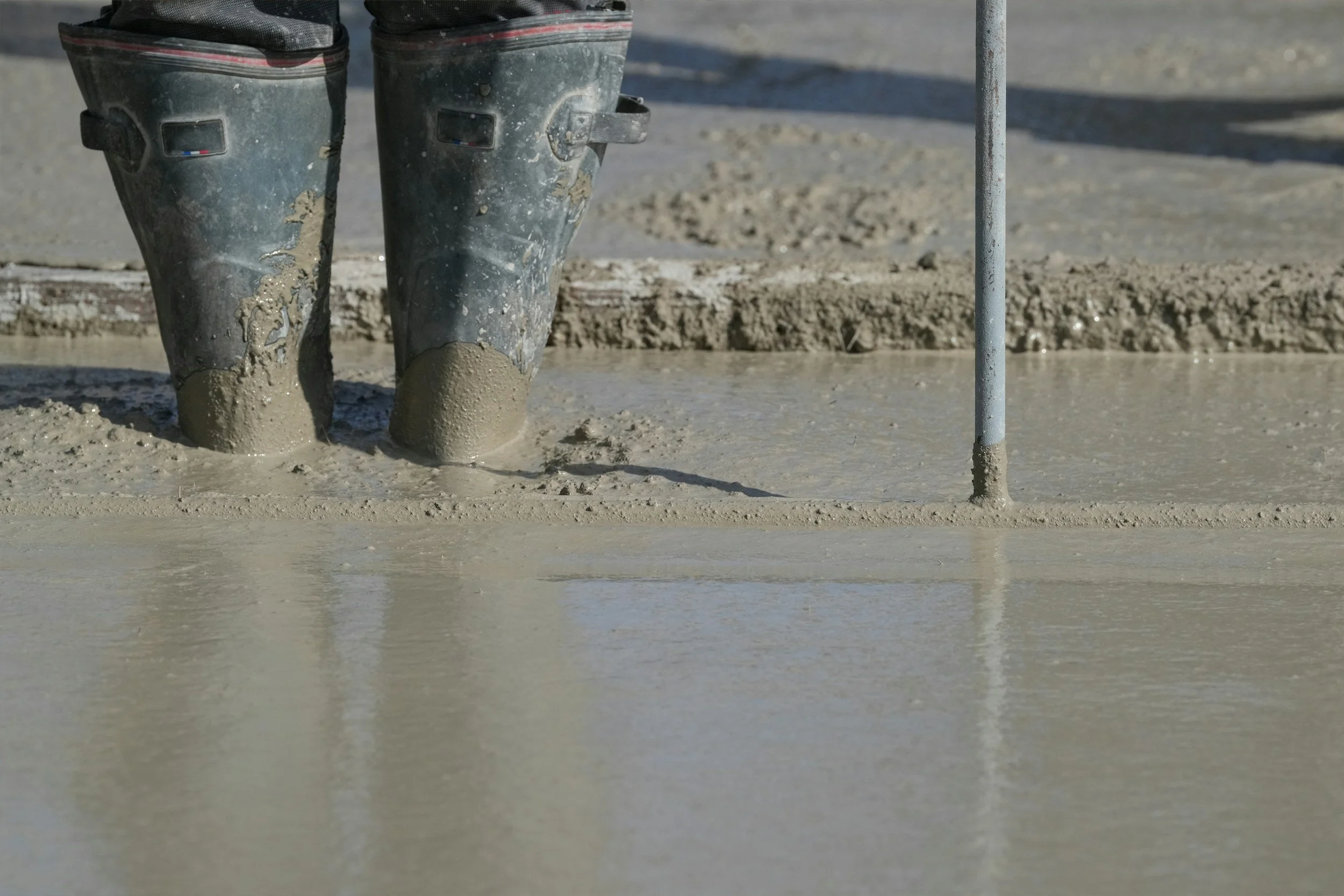 Close-up of black rubber boots with mud on them standing on wet concrete, with a metal pole nearby, indicating a construction or roadwork site.