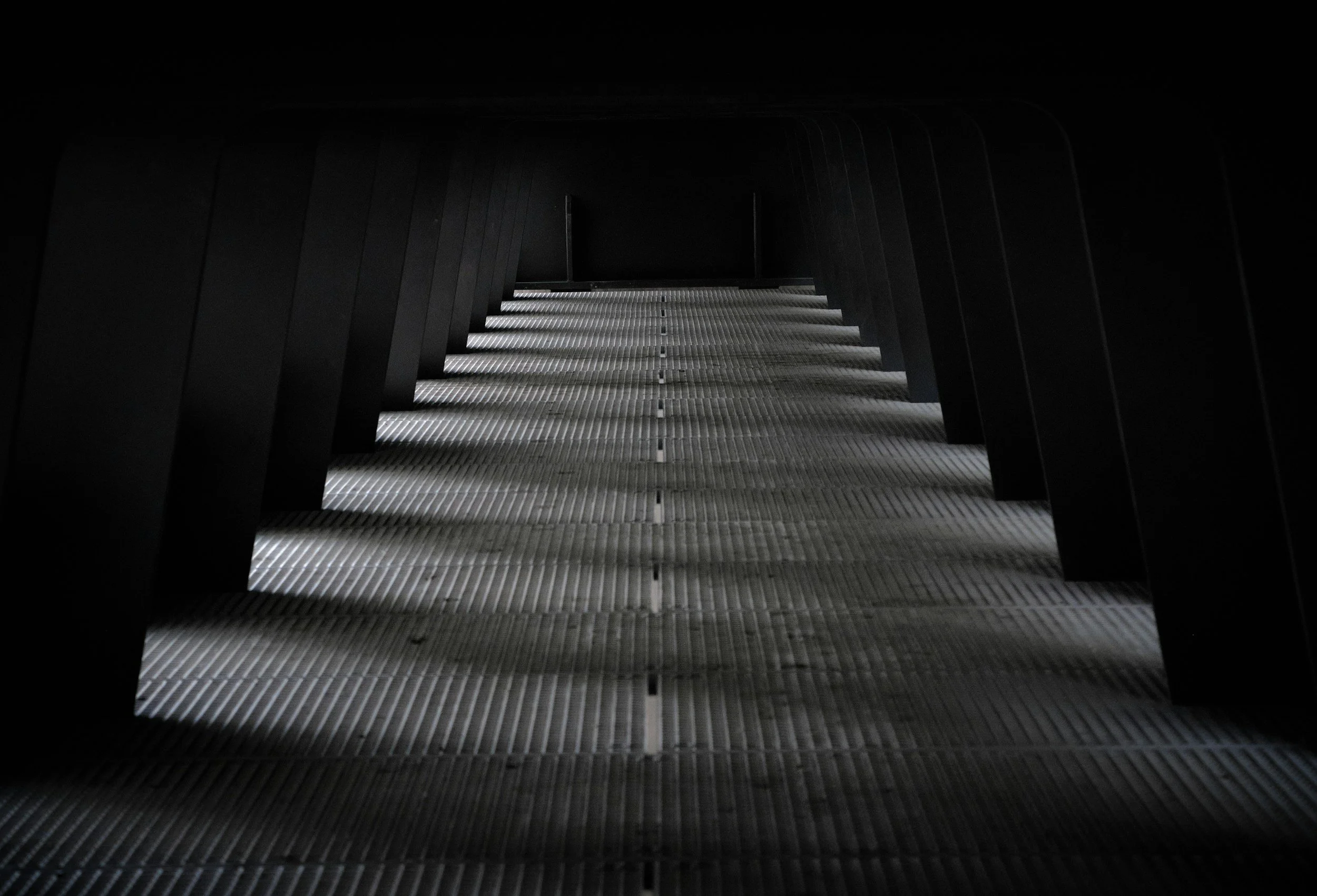 Dimly lit underground passageway with metallic grates on the floor, curved black walls, and minimal lighting at the end of the corridor.