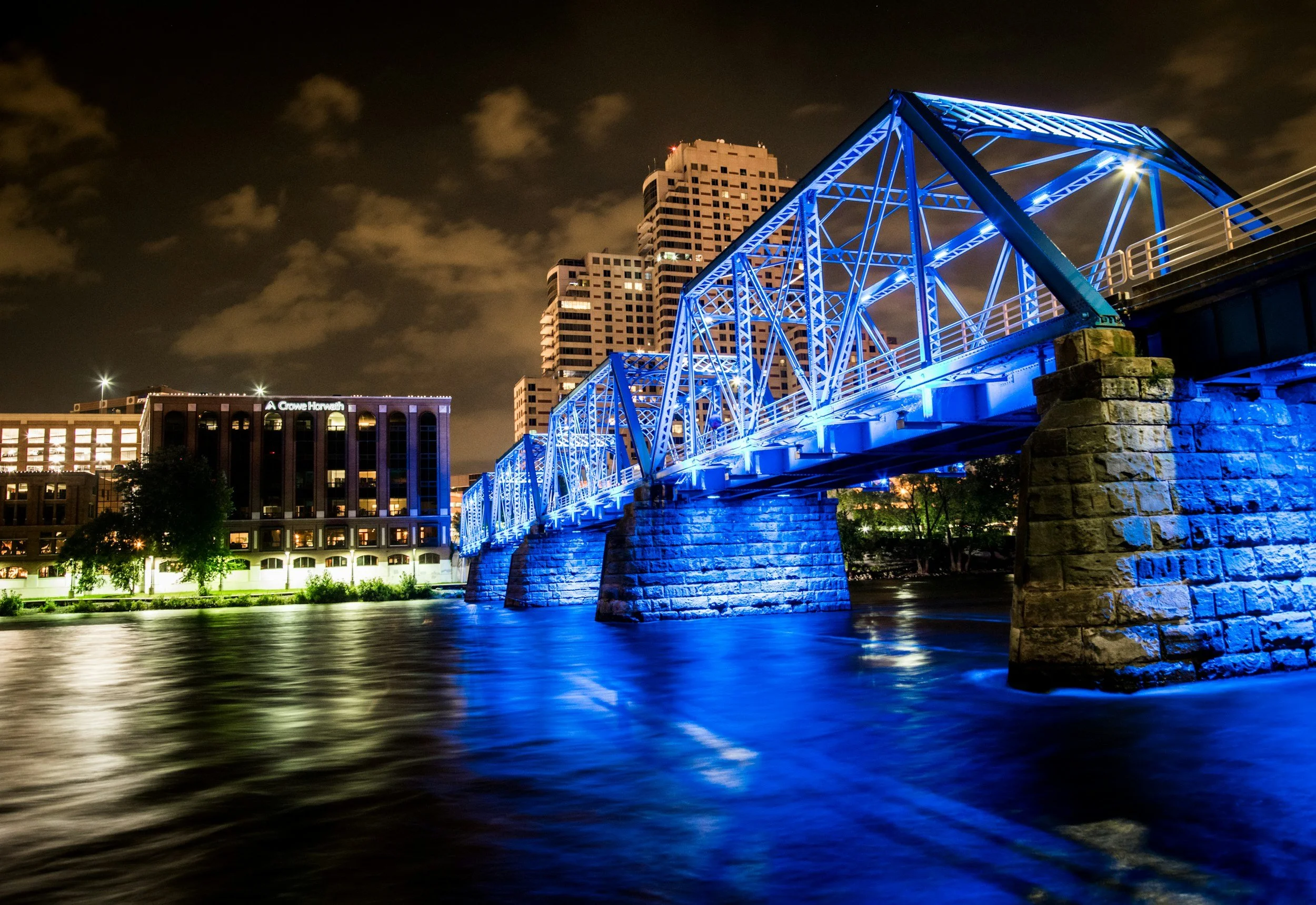 Nighttime view of a blue lit bridge over a river with city buildings in the background.