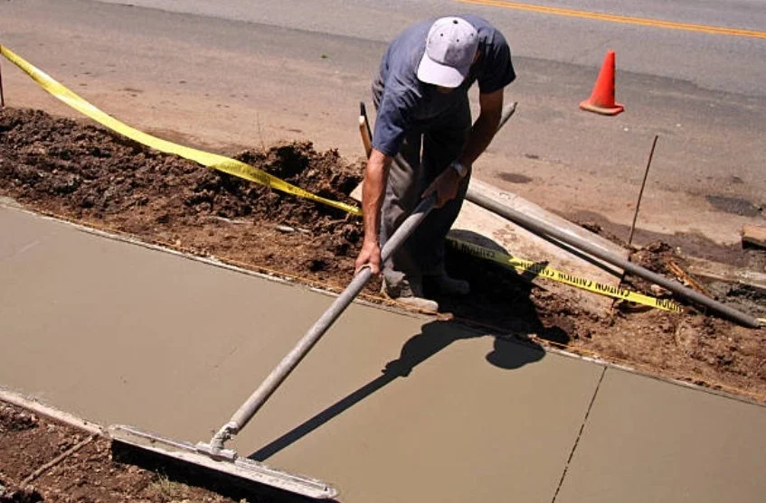 A construction worker with a cap smoothing wet concrete on a sidewalk with a long-handled tool, yellow caution tape, orange traffic cones, and a paved road in the background.