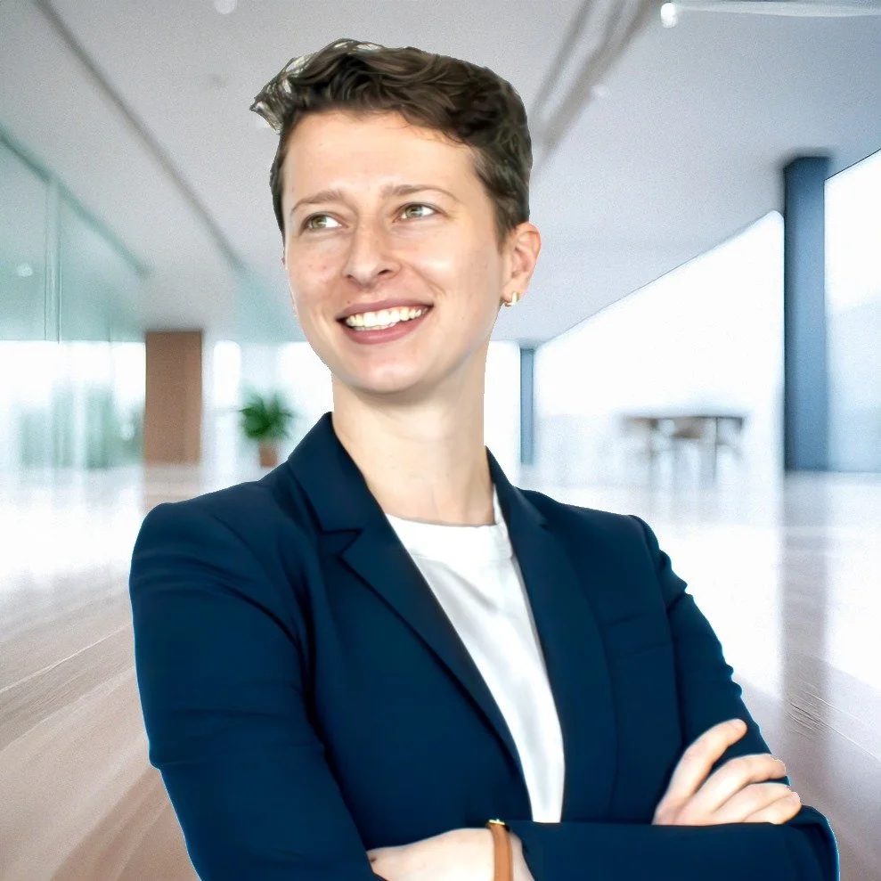 A professional woman with short dark hair wearing a navy blazer and white shirt, smiling confidently in a modern office building.