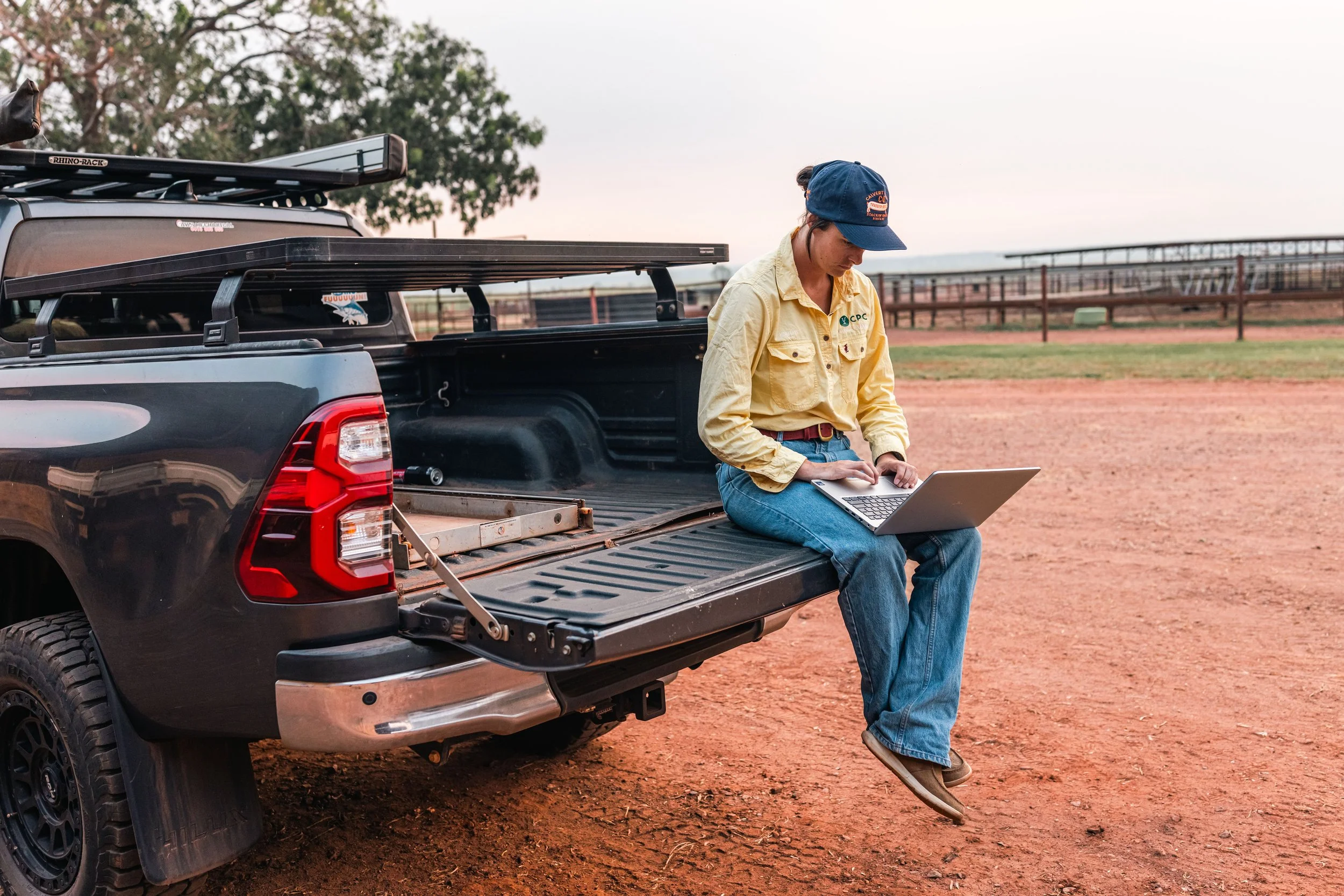 A woman in a yellow shirt, blue jeans, and a blue cap sits on the tailgate of a black pickup truck on a dirt field, working on a laptop with a sports stadium or arena in the background.