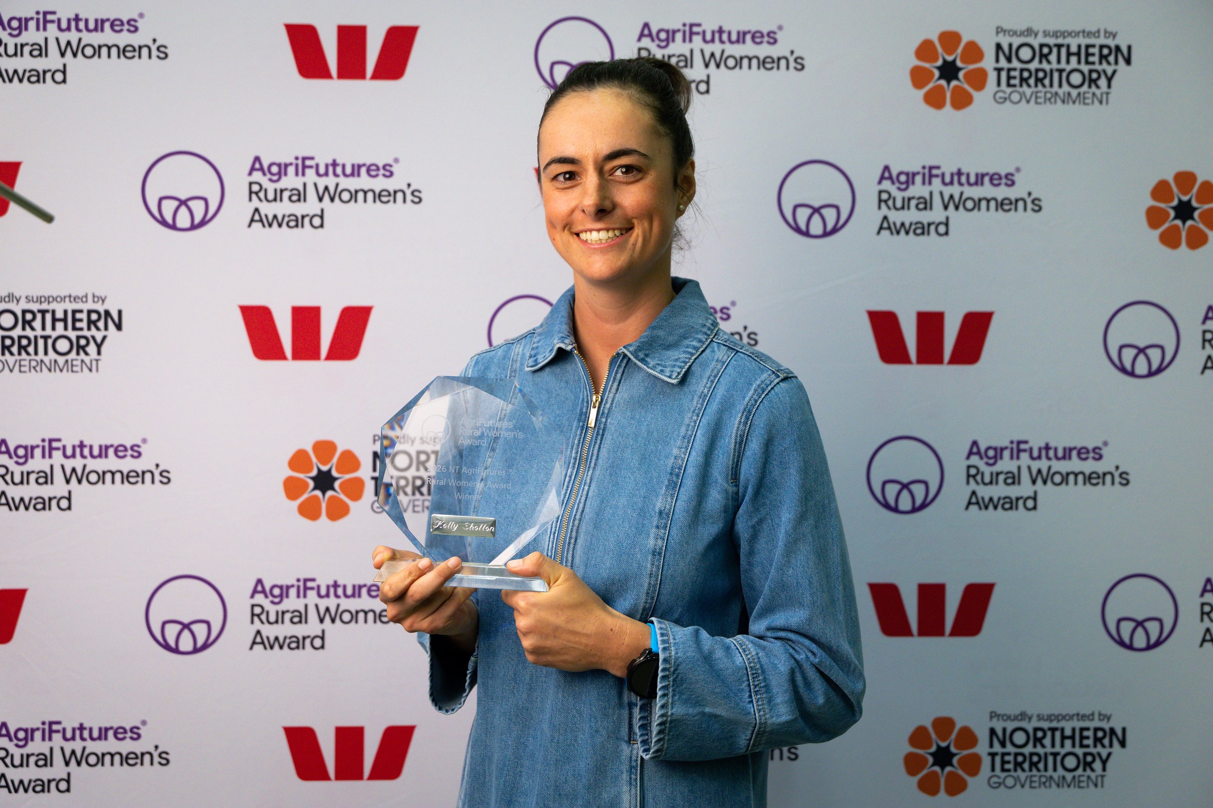 A woman smiling and holding a glass award at the AgriFutures Rural Women’s Award event, standing in front of a backdrop with logos from AgriFutures and the Northern Territory Government.
