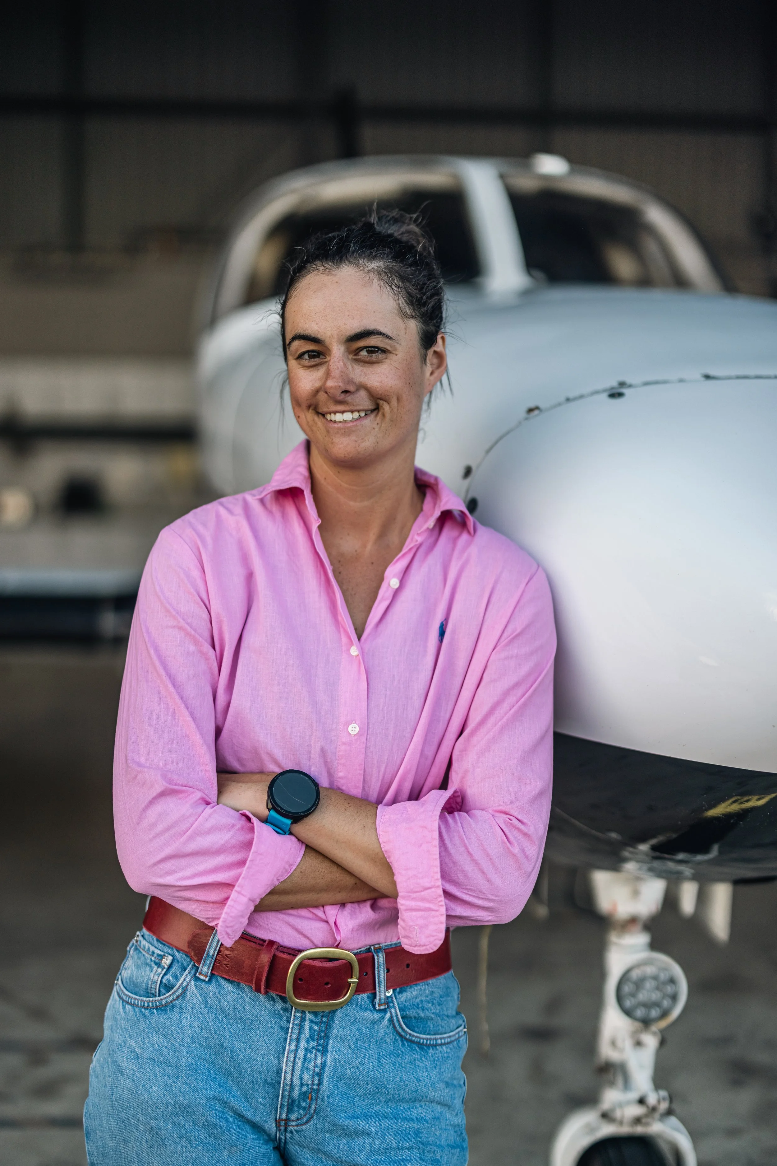 A woman standing with arms crossed, smiling, in front of a small airplane inside a hangar, wearing a pink shirt, blue jeans, and a watch on her left wrist.