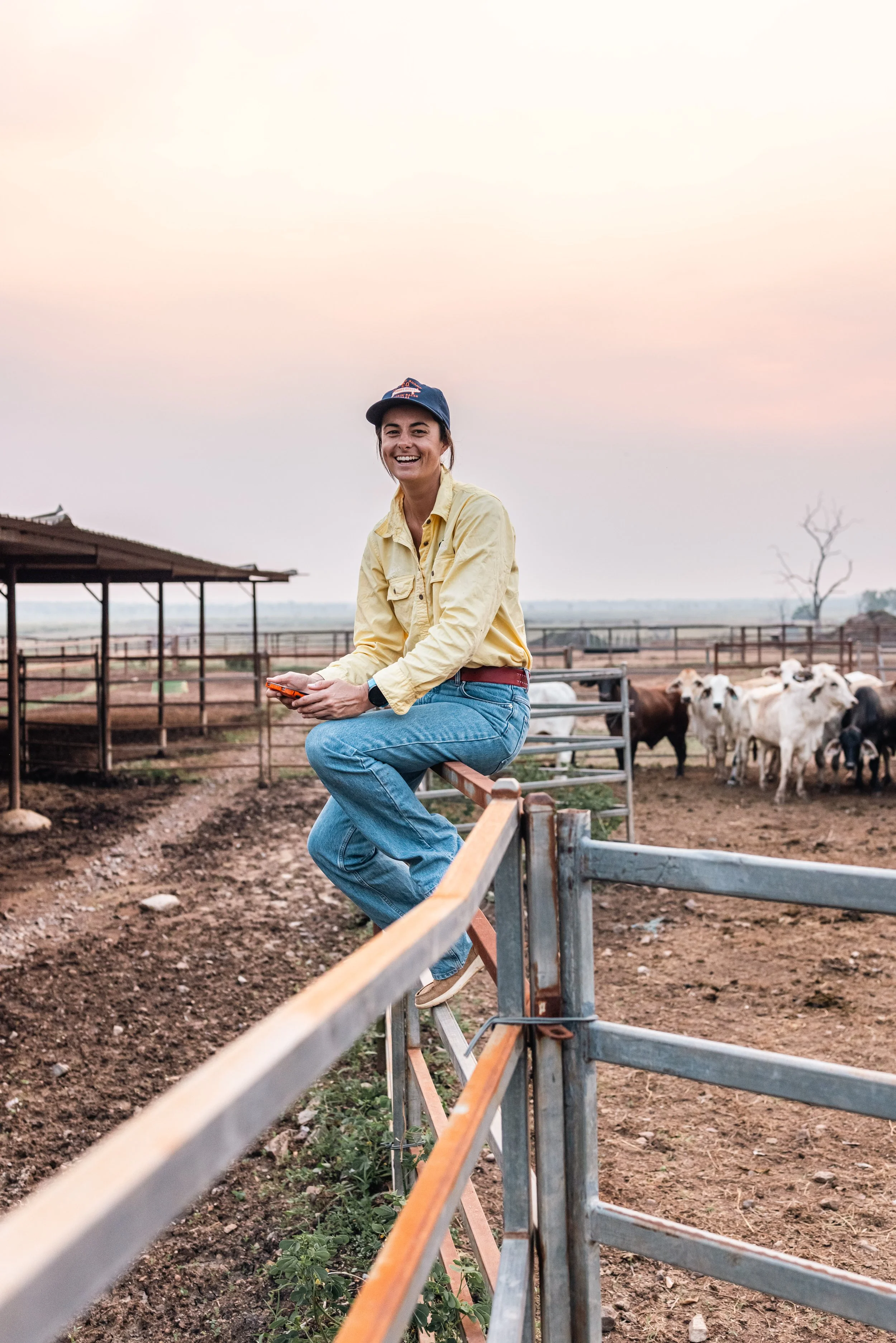 A woman in a yellow jacket, blue jeans, and a blue cap sitting on a fence at a farm, smiling with cows in the background during sunset.