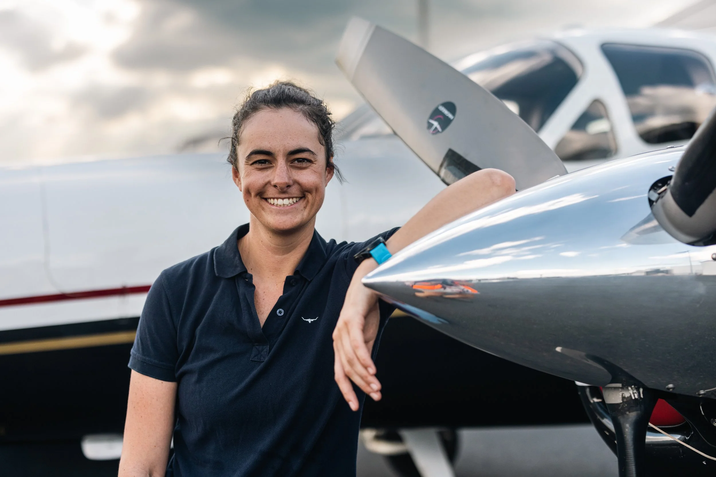 Young woman with dark hair tied back, smiling, standing outside next to a small aircraft, possibly a sailplane or glider, with other airplanes visible in the background.