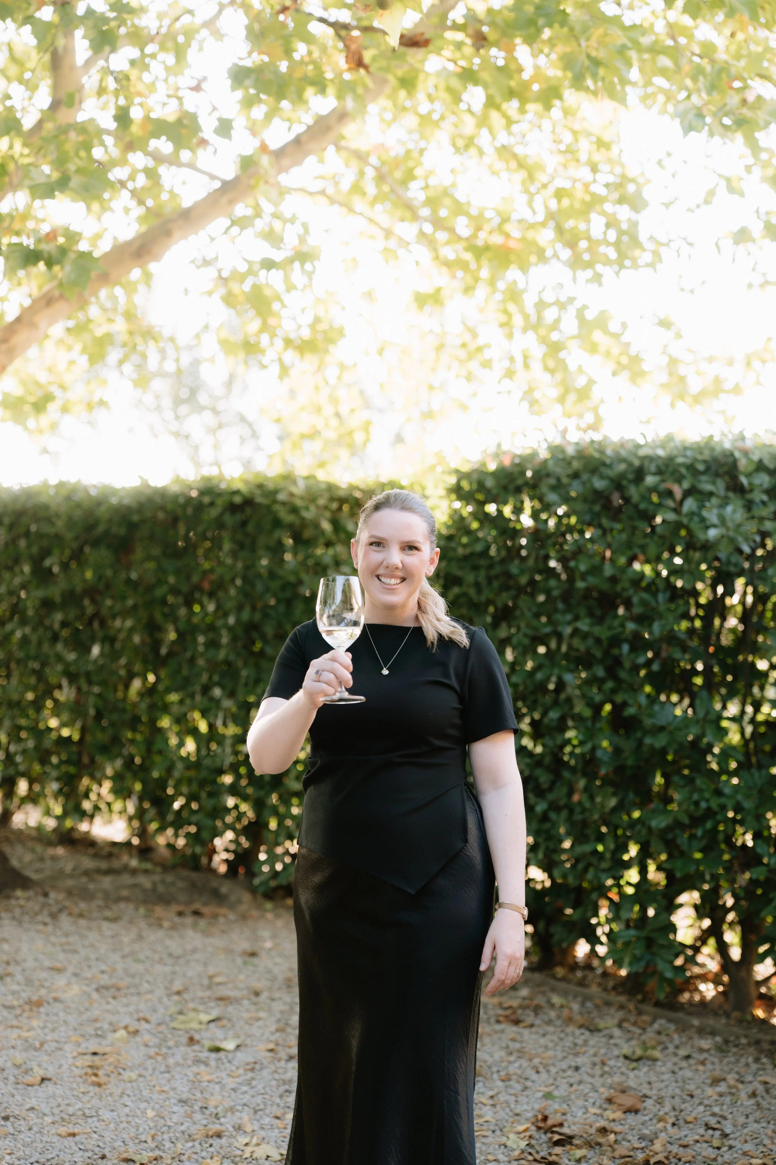A woman in a black dress holding a wine glass outdoors with green foliage and trees in the background.