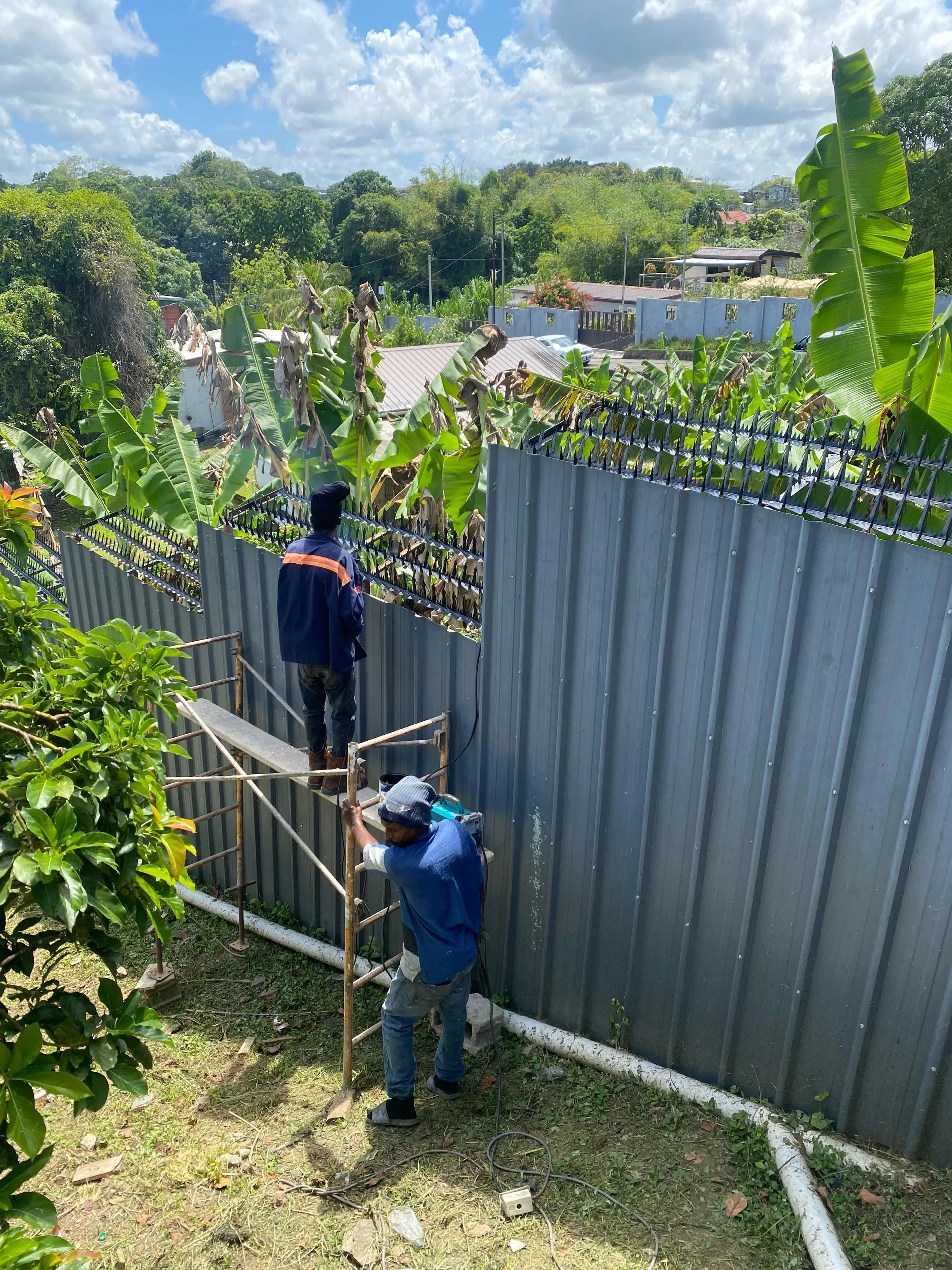 Two workers are installing or repairing a corrugated metal fence on a grassy area with trees and foliage nearby. One worker is standing on a scaffold and the other is on the ground, both working on the fence. There are electrical cords and a power to