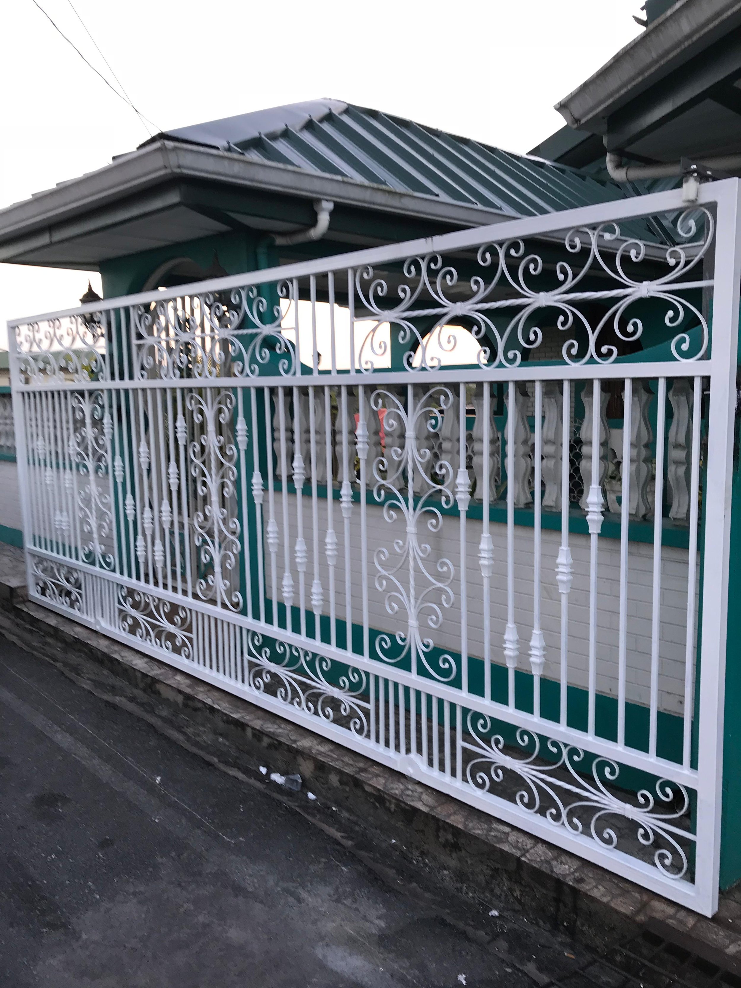 A white decorative metal gate with intricate scrollwork designs, installed in front of a house with a teal exterior, a metal roof, and visible window shutter.