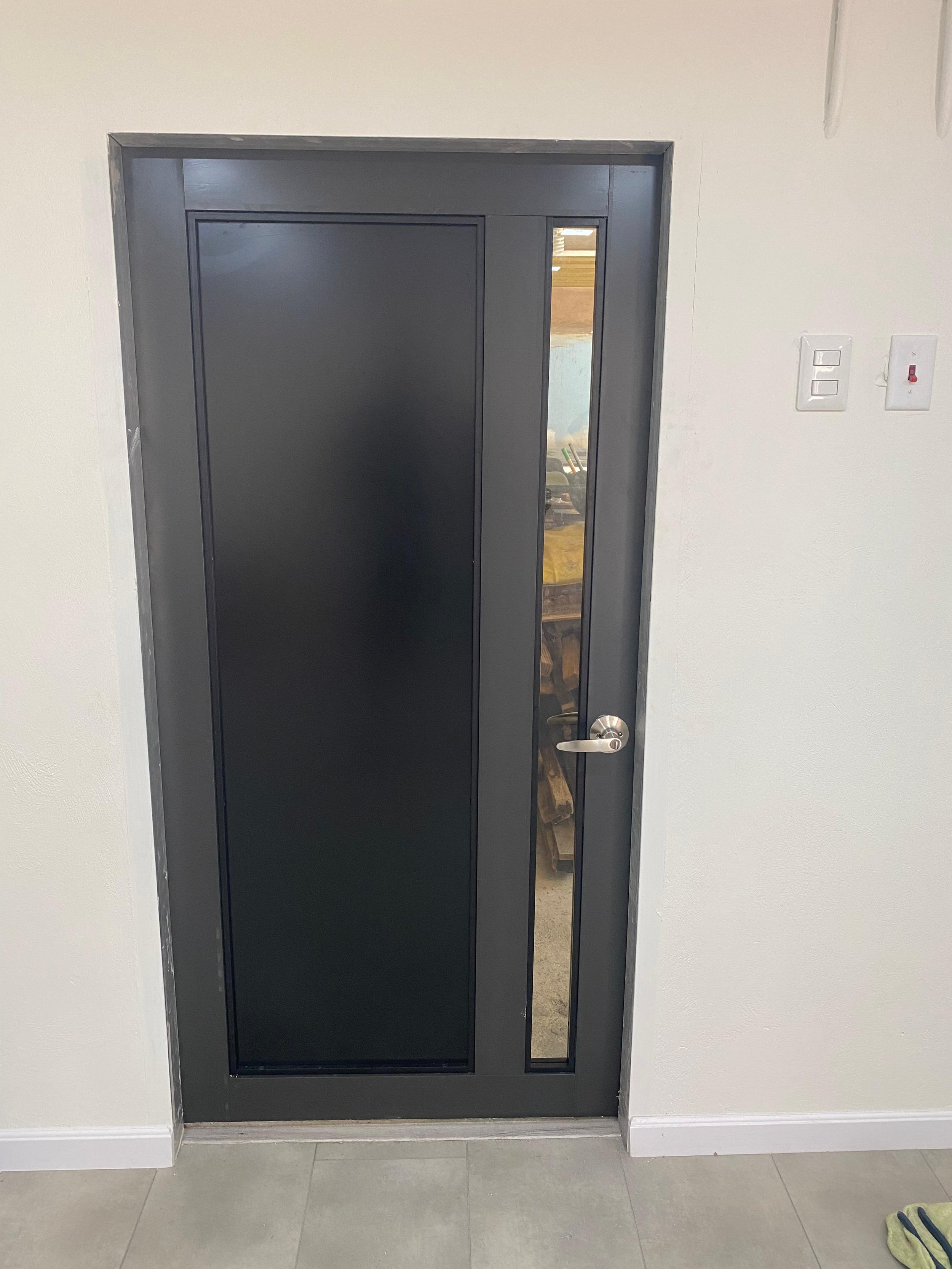 A black security door with a small vertical window on the right side, mounted on a white wall. There are two light switches on the wall to the right of the door. Through the window, a room with a brown couch and some boxes or storage items can be see