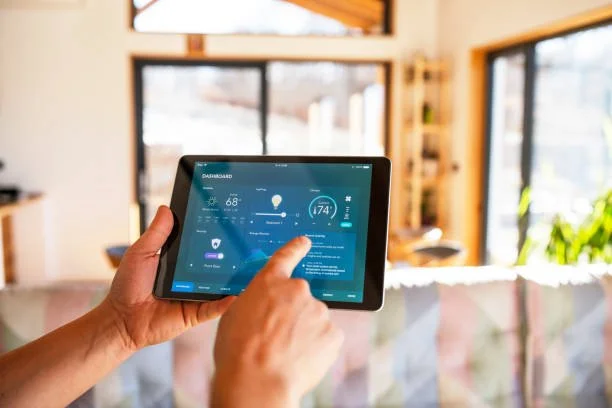 A person is holding a tablet showing a smart home dashboard in a sunlit living room with large windows and a sofa.