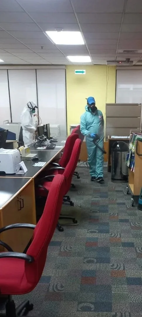 Two people in full personal protective equipment, including suits, masks, and gloves, cleaning and disinfecting an office space with desks, chairs, and office supplies.
