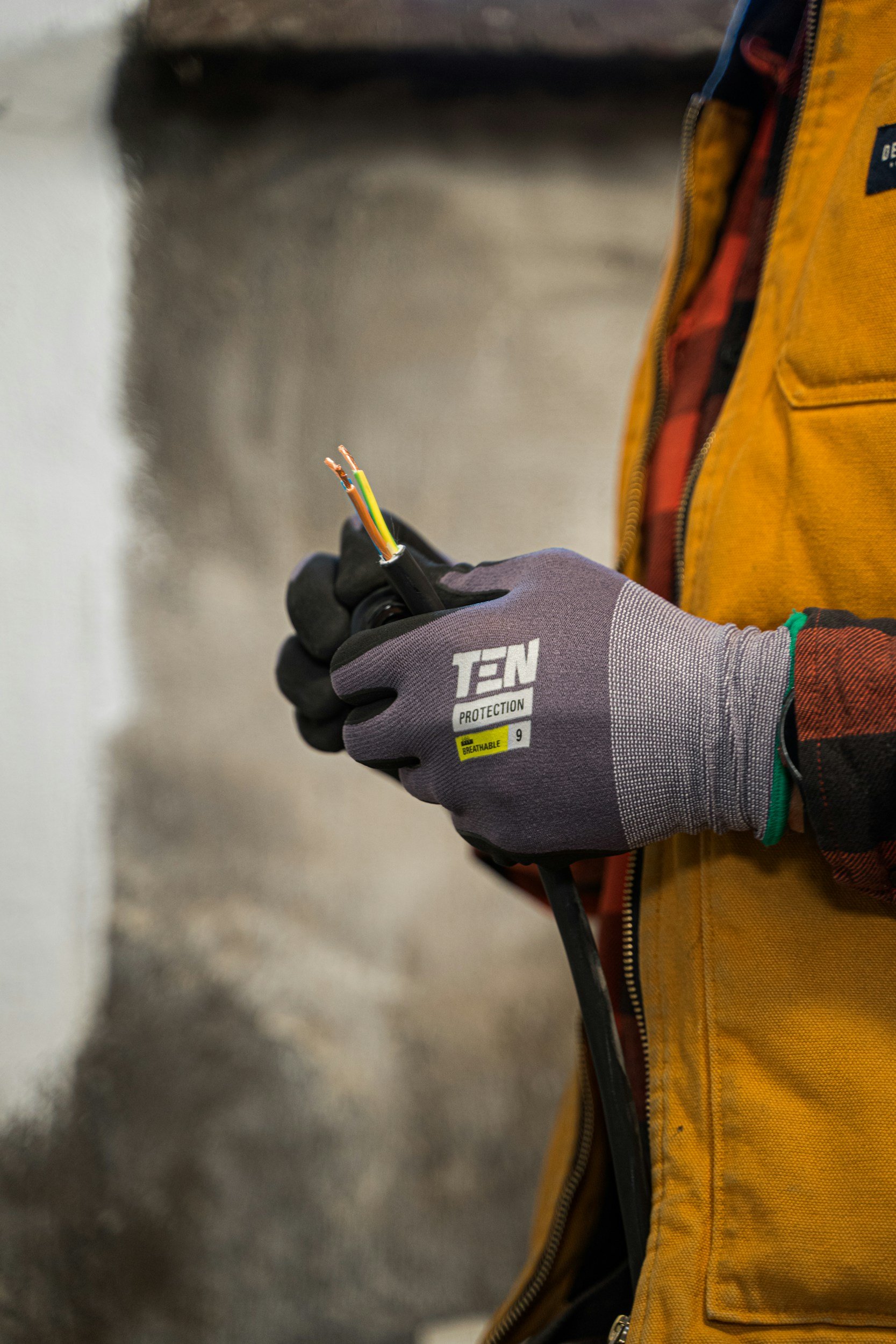 A person wearing a gray glove holding a black electrical wire with multiple colored wires exposed at the cut end, in front of a person in a yellow jacket.