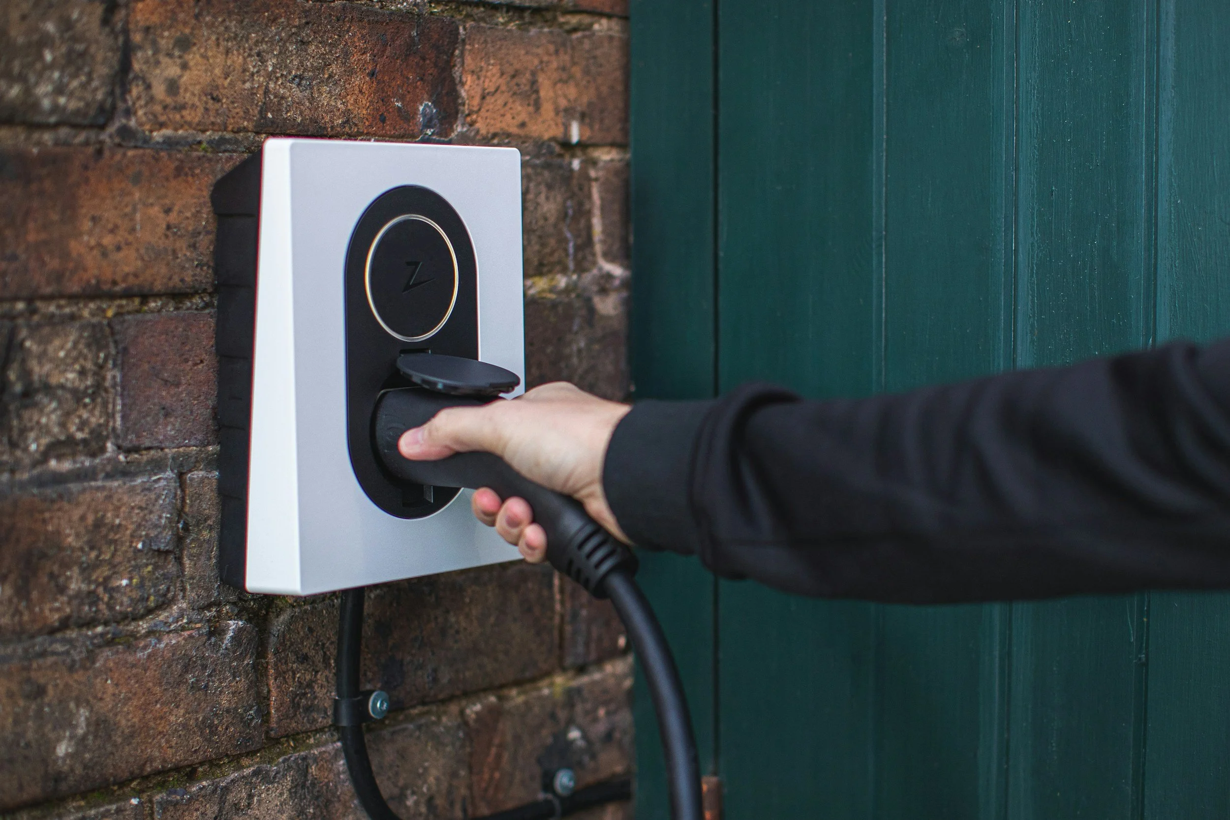 A person is plugging an electric vehicle into a wall-mounted charging station outside next to a brick wall and a green wooden door.