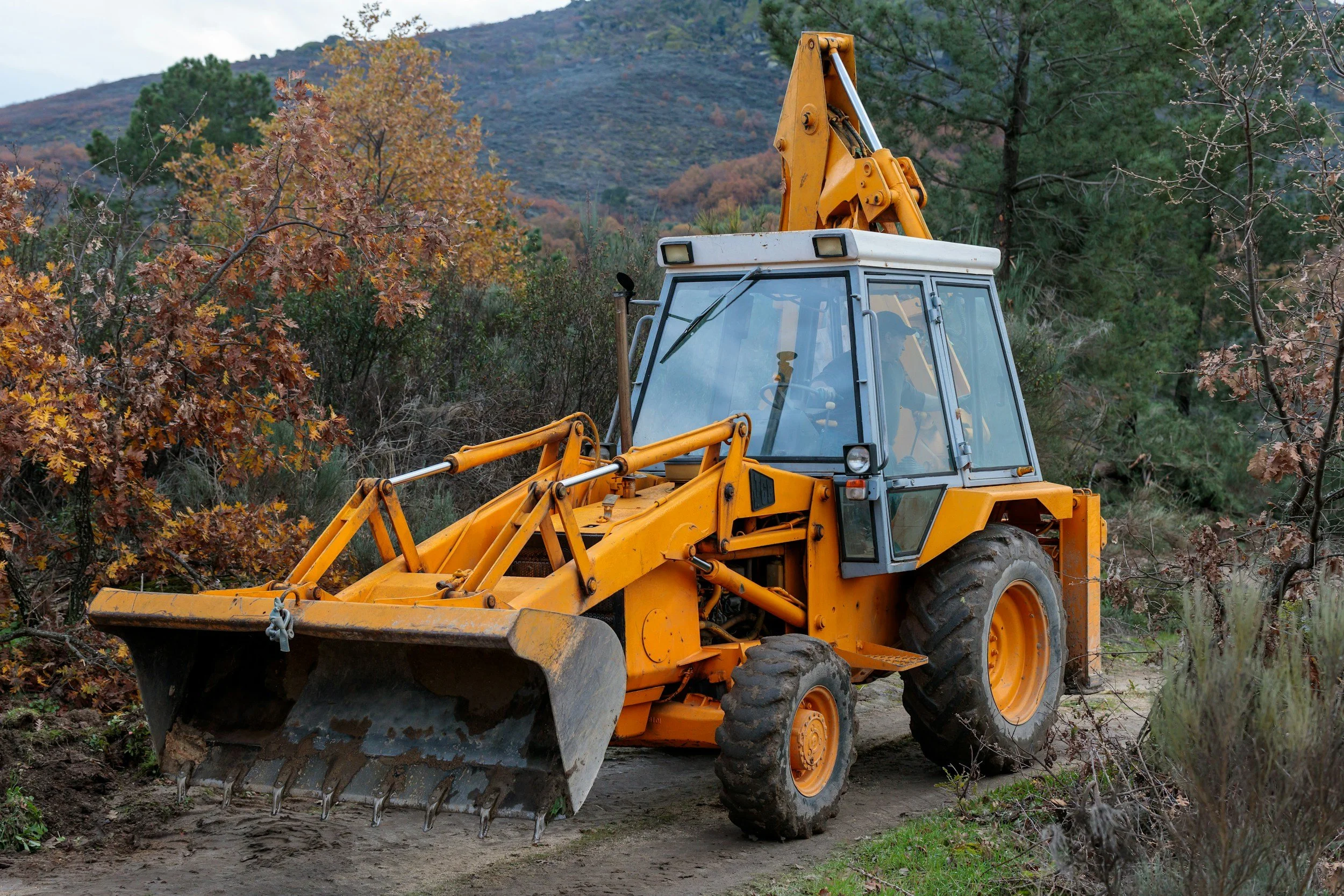 Yellow bulldozer clearing trees and soil in a forested area with mountains in the background.