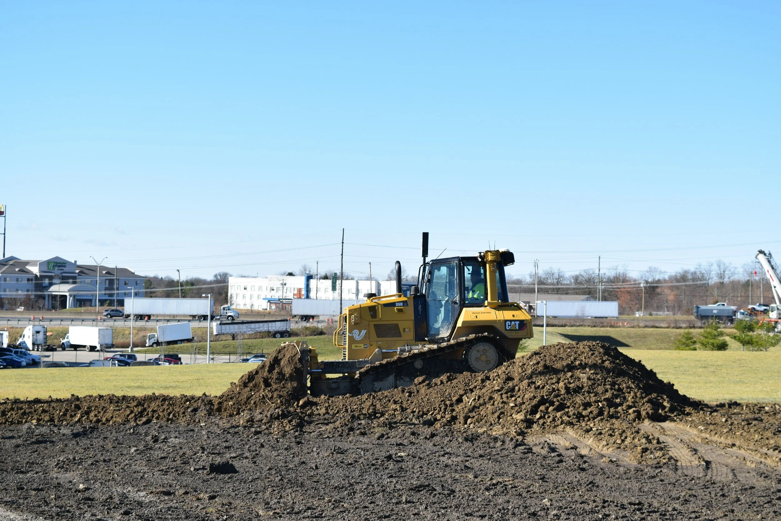 A yellow bulldozer working on a construction site with a mound of earth in the foreground. In the background, there are buildings, traffic, and electrical poles under a clear blue sky.