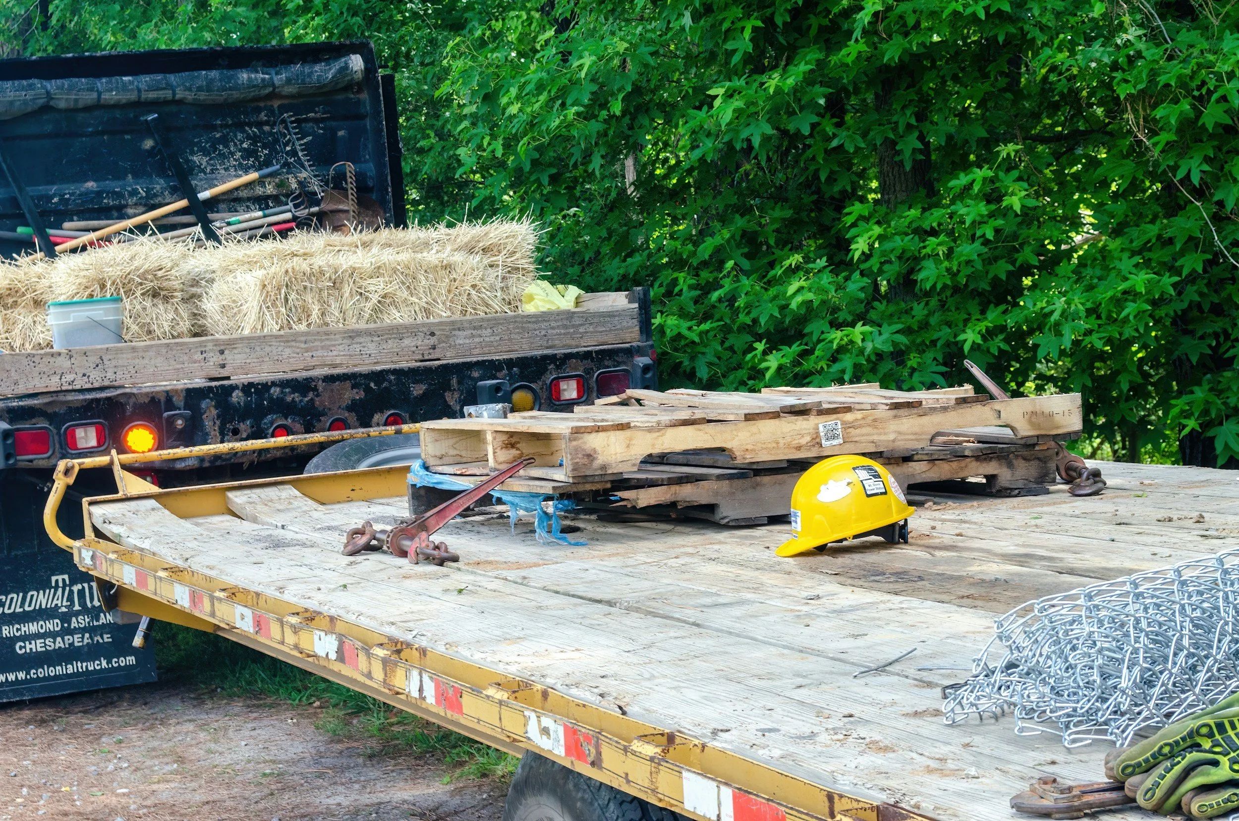 Flatbed truck carrying hay bales, tools, and construction safety gear parked outdoors near green trees.