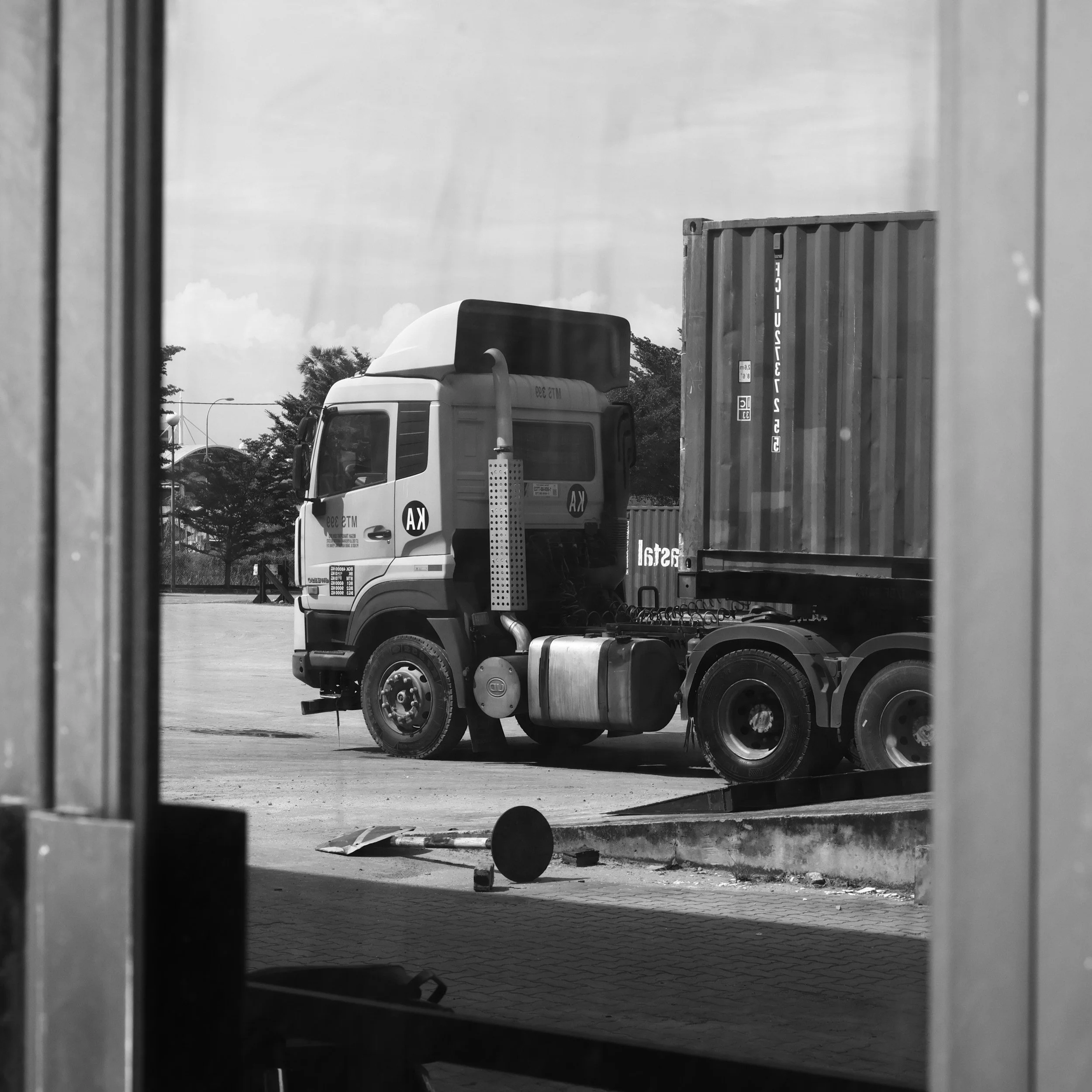 View of a parking lot with a large white semi-truck carrying a shipping container, seen through an opening between two vertical structures, with trees and cloudy sky in the background.