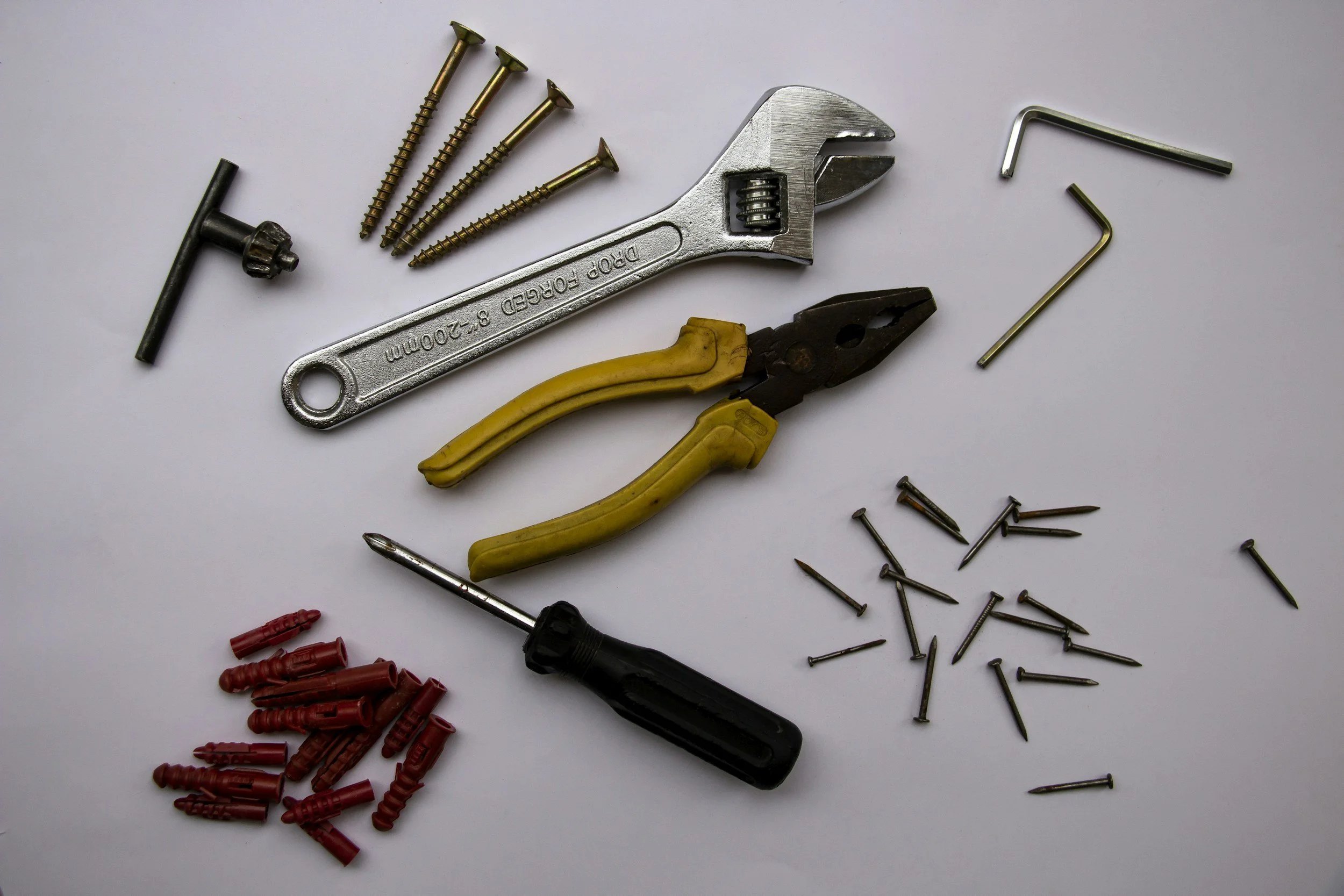 Assorted hardware tools and parts including a wrench, pliers, screws, nails, and wire connectors on a white surface.
