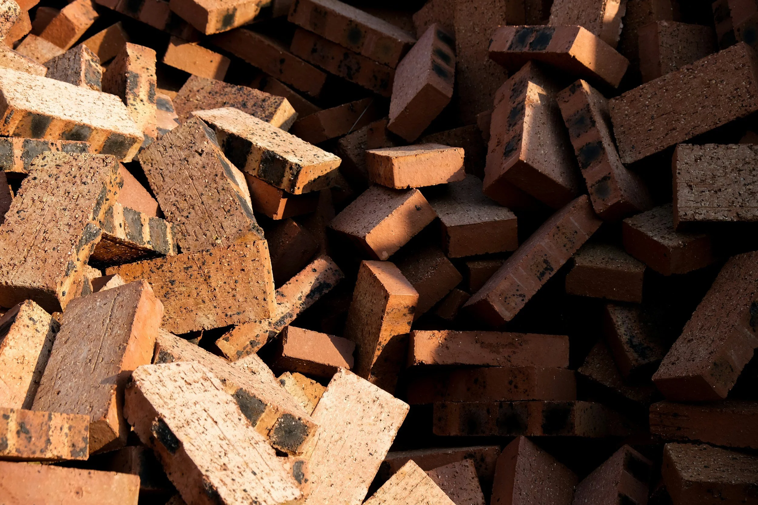 Stack of red bricks with dark spots, some bricks have chipped edges, sunlight illuminating the bricks creating shadows.