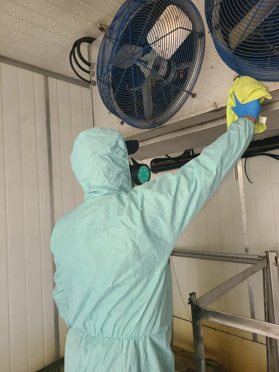 A person wearing a full protective suit, gloves, and a mask cleaning a large industrial fan with a yellow cloth.