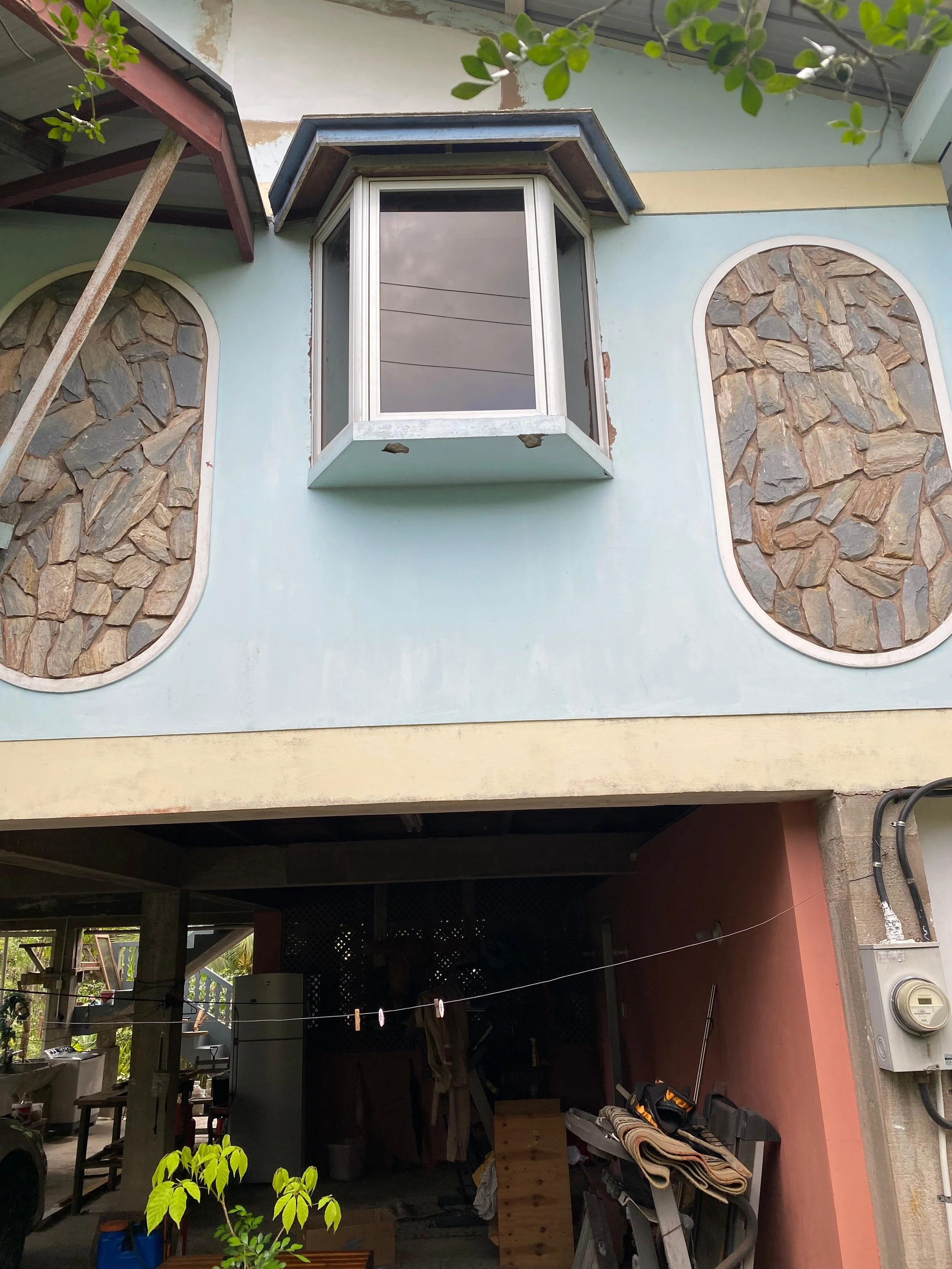 The photo shows the exterior of a building with a light blue wall that has decorative stone insets framed in white. There is a bay window protruding from the wall with a gray sloped roof above it. The lower part of the building appears to be an open 