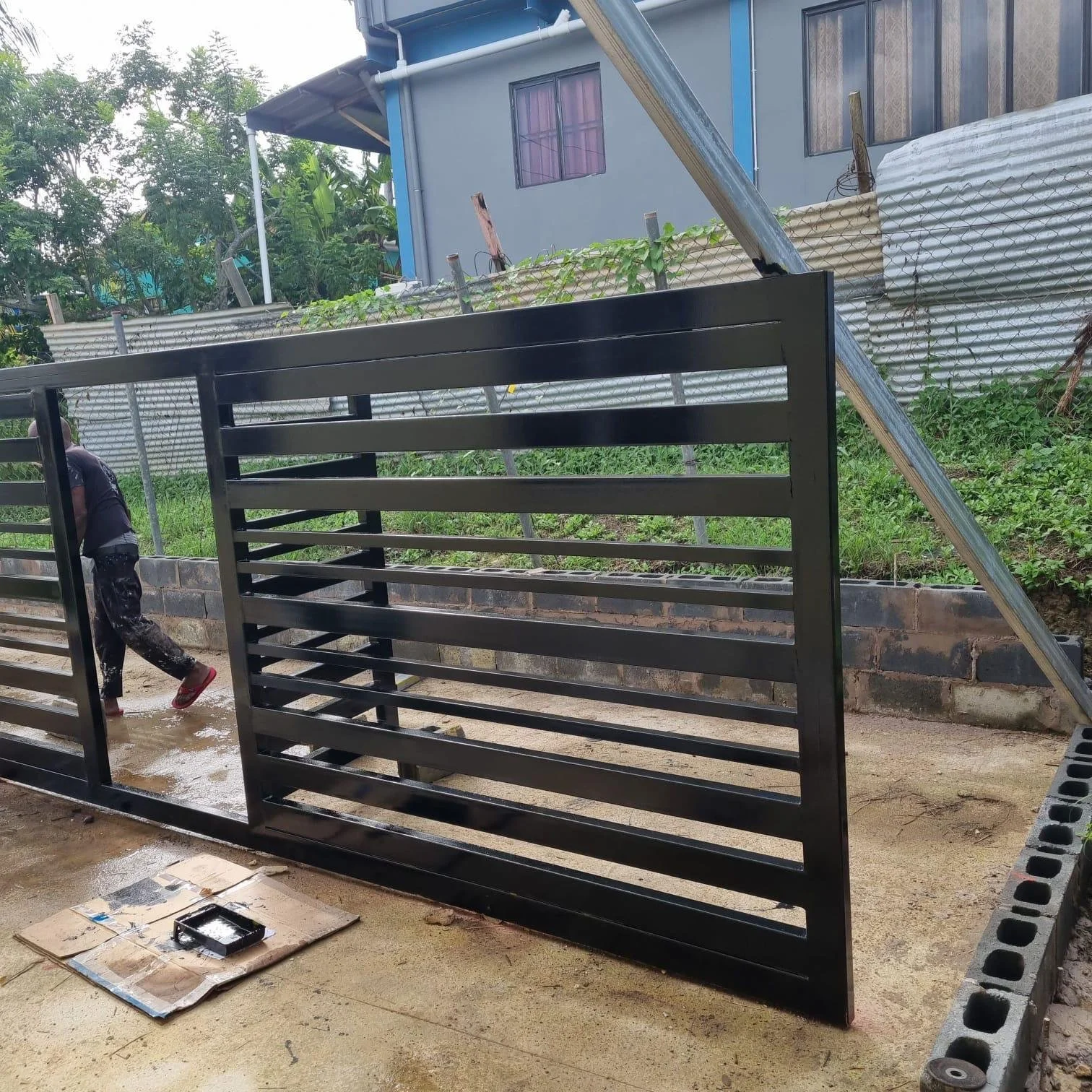 A person working outside, painting a black metal gate, with painting supplies on a cardboard on the ground, and a house with purple curtains and a grey exterior wall in the background.
