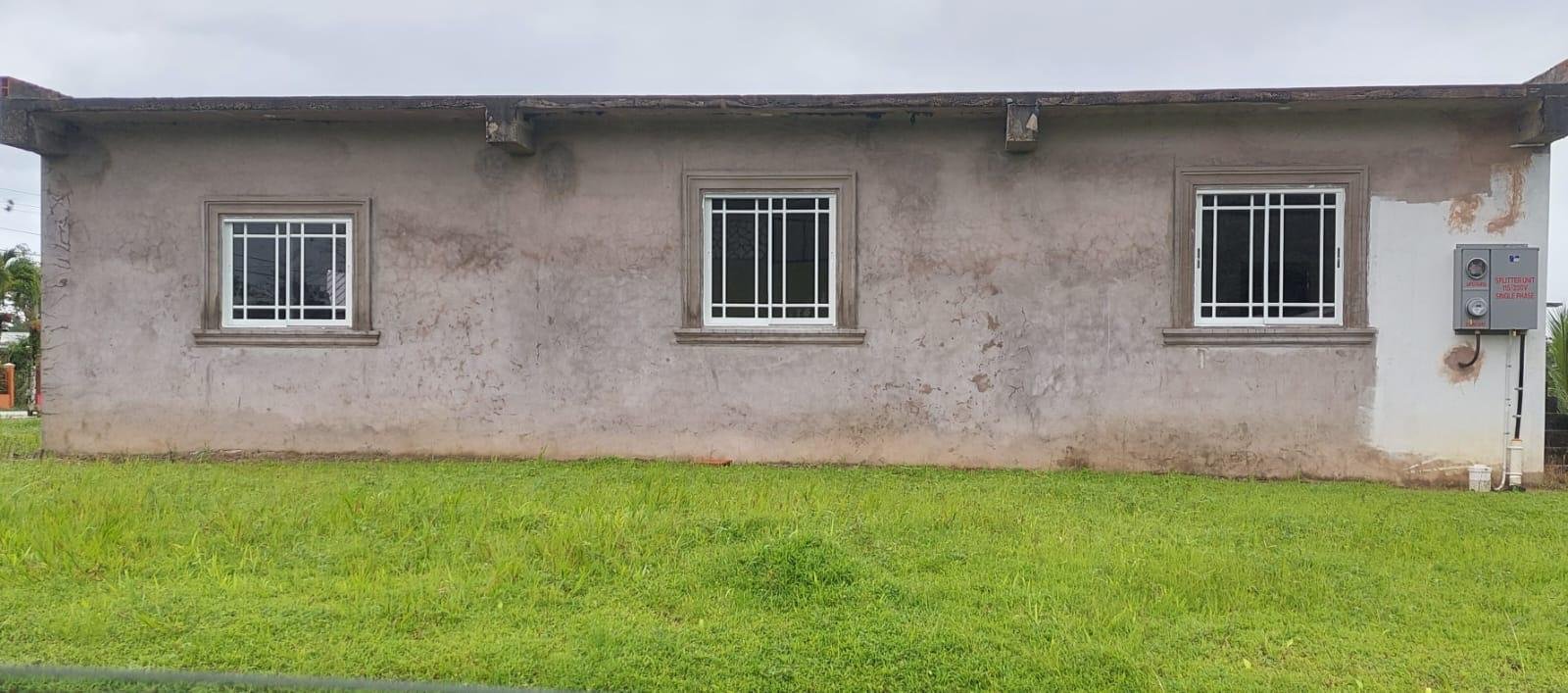 A plain, single-story house with three windows and a gray exterior wall. The house has a slightly weathered appearance and a grassy lawn in front. An electrical panel is mounted on the right side of the wall.