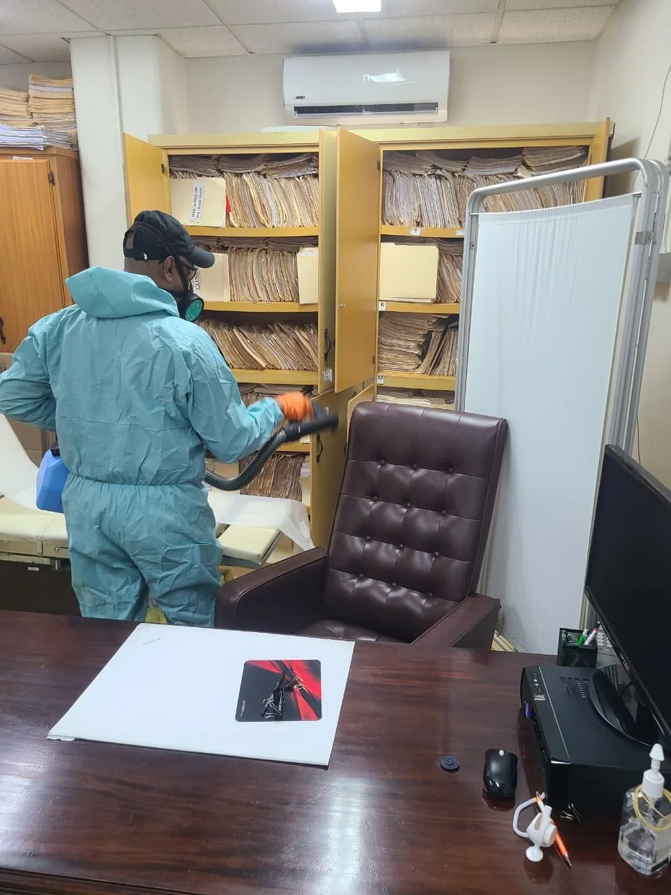 Person in a blue protective suit and mask using a vacuum cleaner to disinfect an office with shelves full of files on the background.