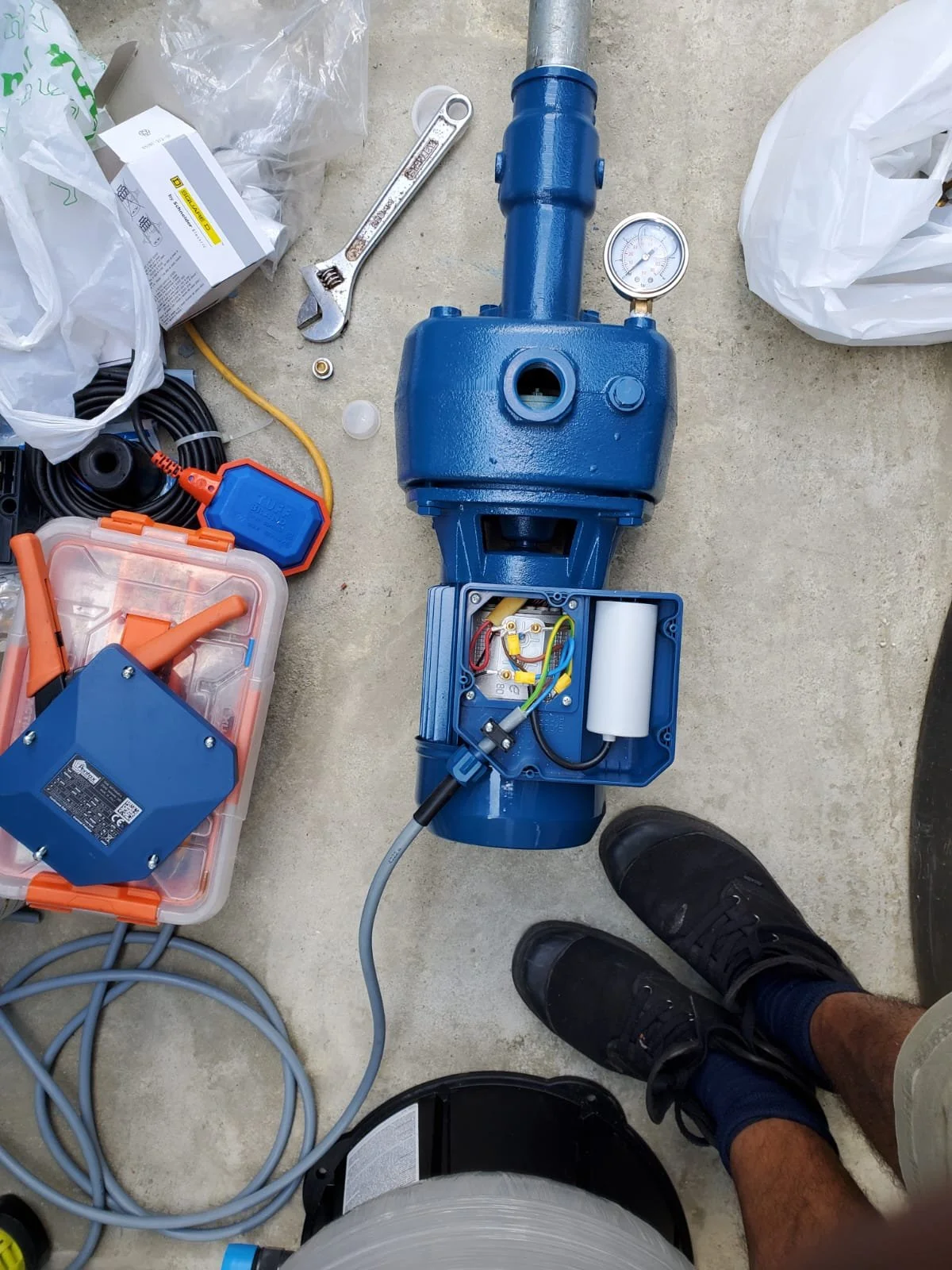 Open blue electrical device with wires and components inside, on concrete floor with tools, cables, and bags around, and a person's feet visible.