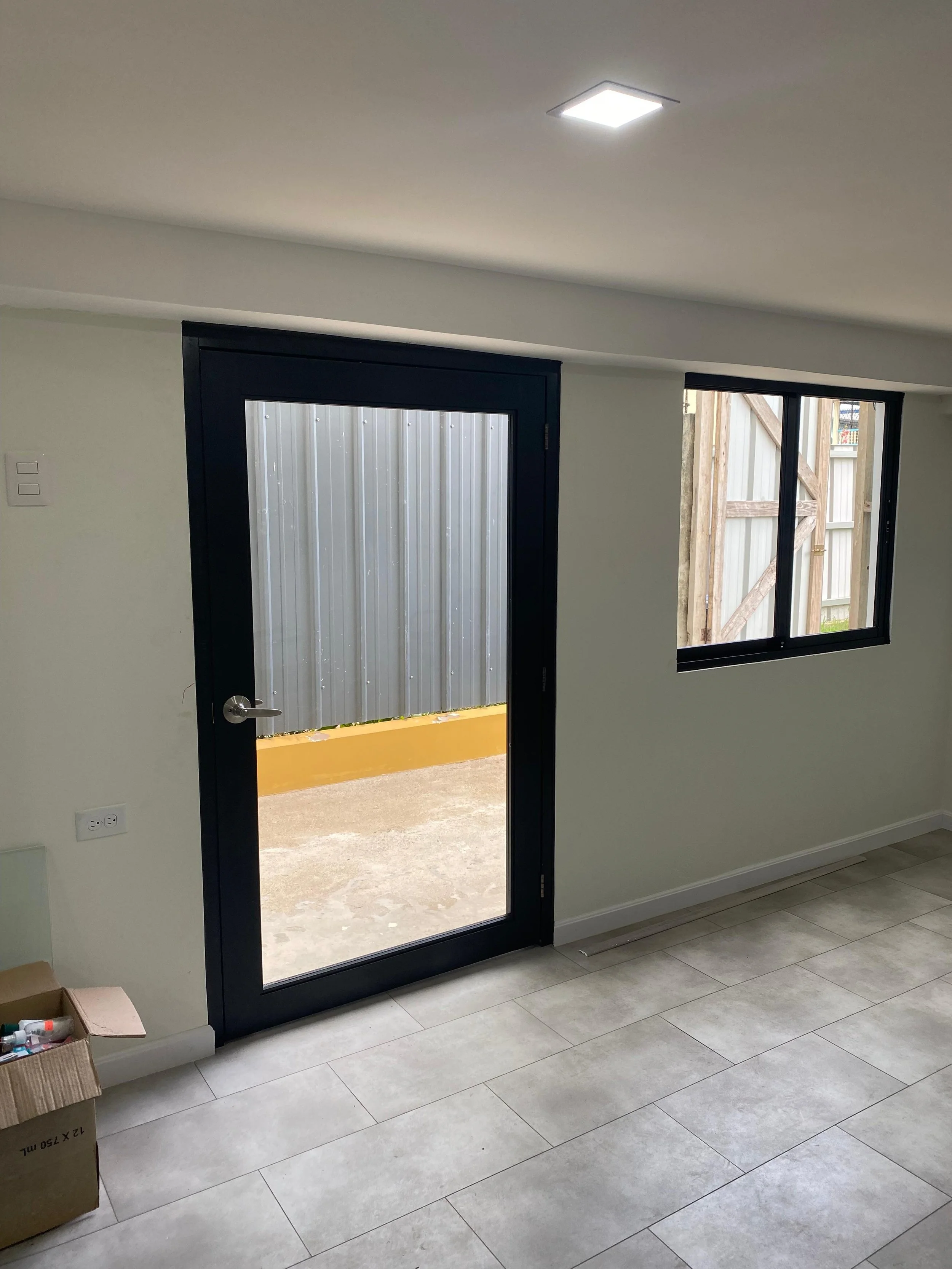 Interior view of a room with a black-framed glass door leading outside to a small yard, and a window showing a wooden fence, with white finished walls and tiled flooring.