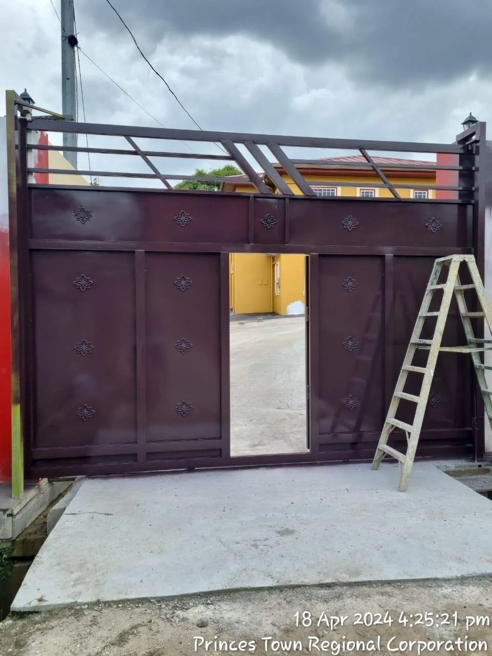 A dark-colored metal gate with decorative patterns, partially open, with a yellow building and cloudy sky in the background. A ladder leans against the right side of the gate.